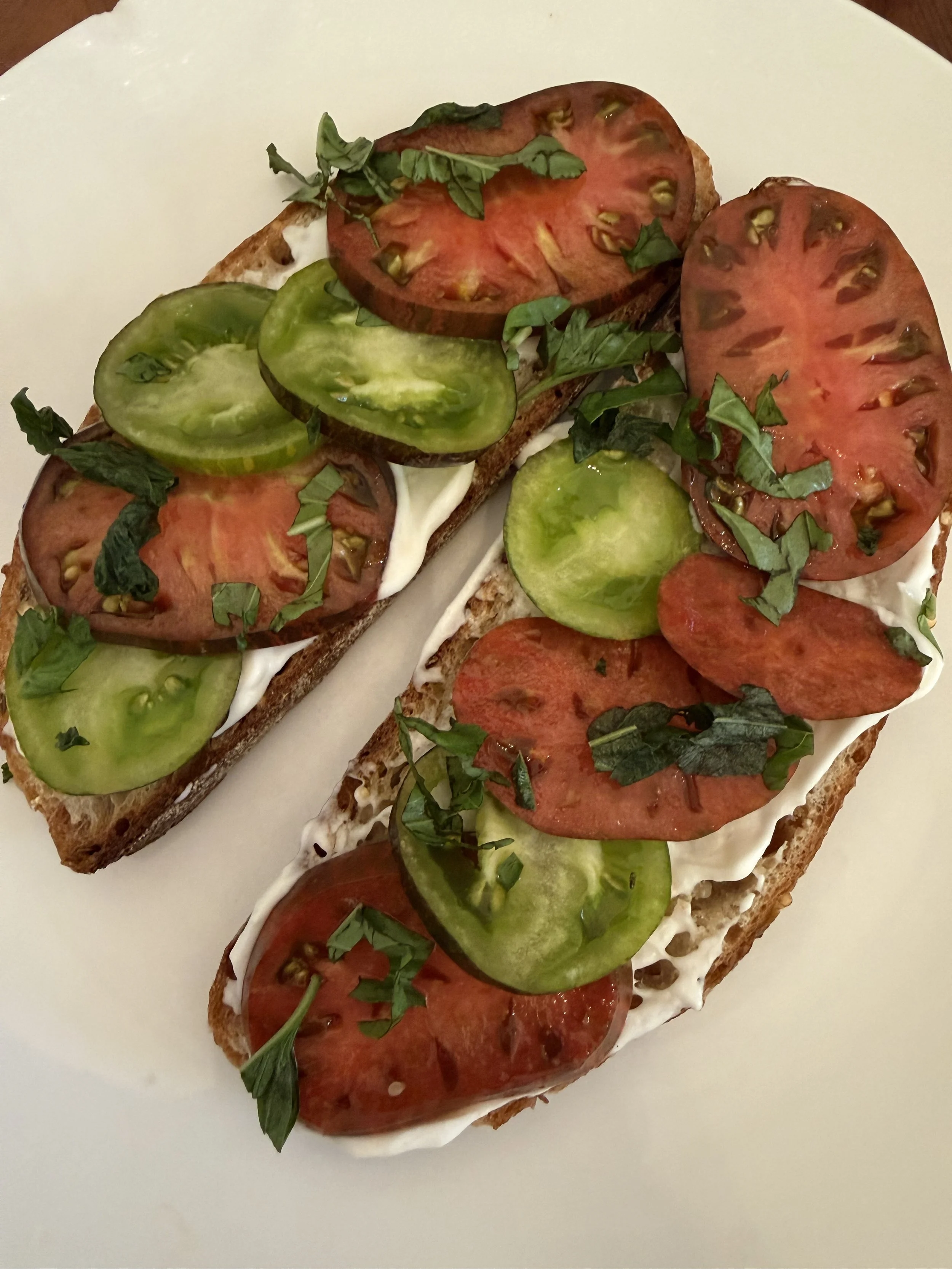 Two slices of toasted bread topped with sliced tomatoes, cucumbers, fresh herbs, and a spread of cream cheese.
