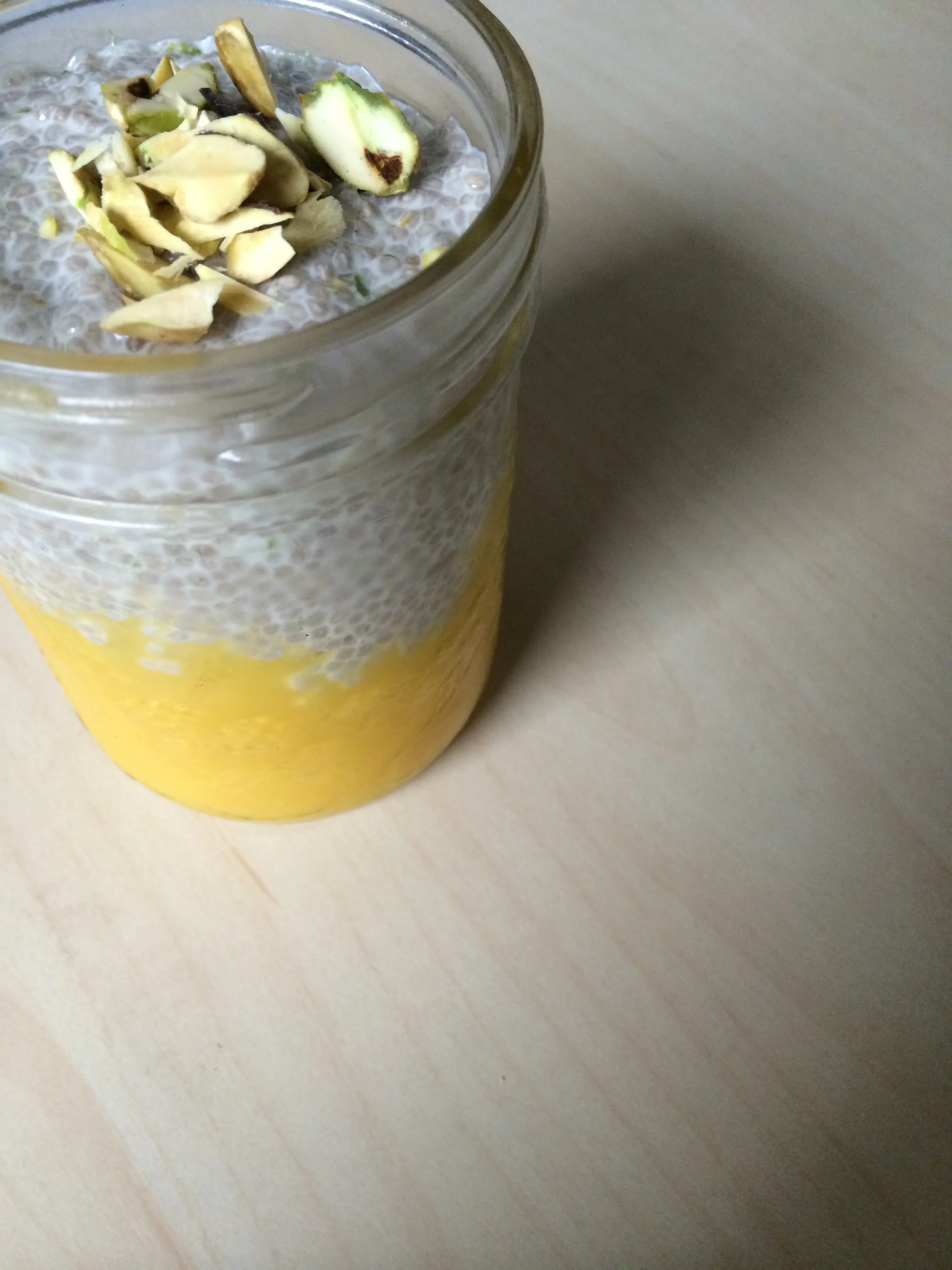 A mason jar filled with a layered dessert or beverage, featuring a yellow bottom layer, a middle layer of gray tapioca pearls or chia seeds, and a top layer of chopped pistachios, all on a light-colored wooden surface.