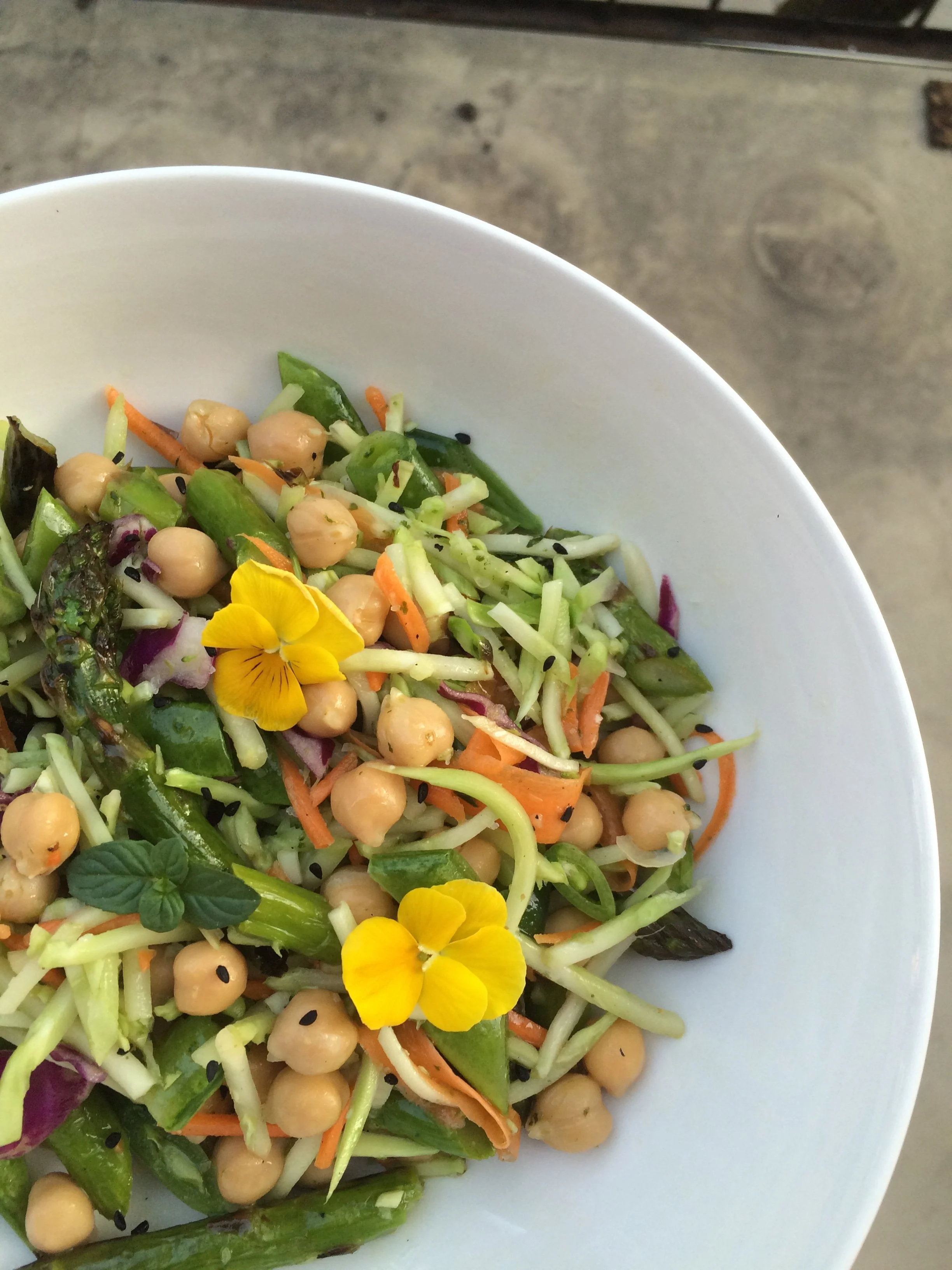 A bowl of mixed salad with chickpeas, shredded carrots, sliced cucumbers, purple cabbage, and edible flowers, garnished with black sesame seeds and mint leaves.