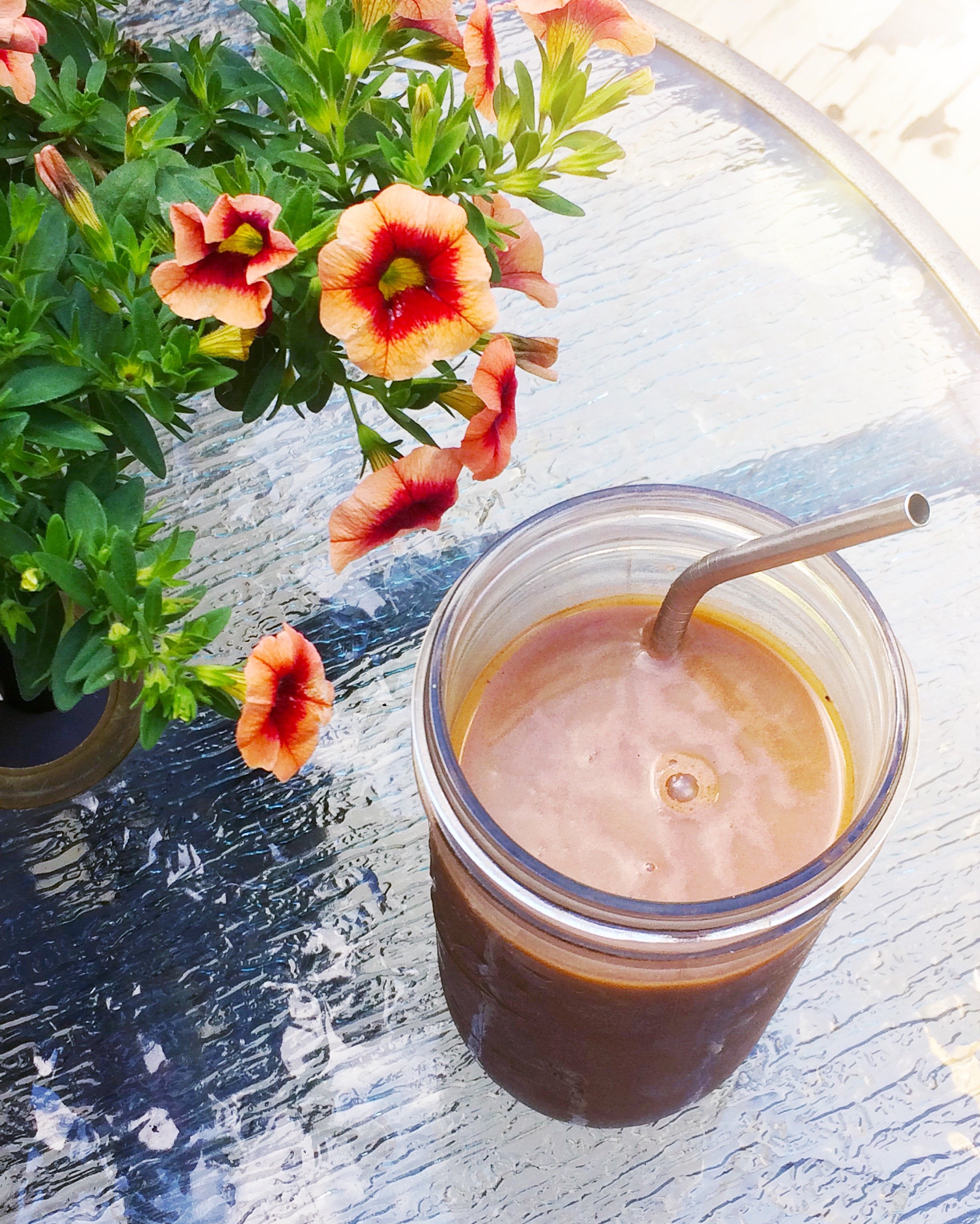 A glass of iced coffee with a metal straw on a textured glass table next to a potted plant with orange and red petunia flowers.