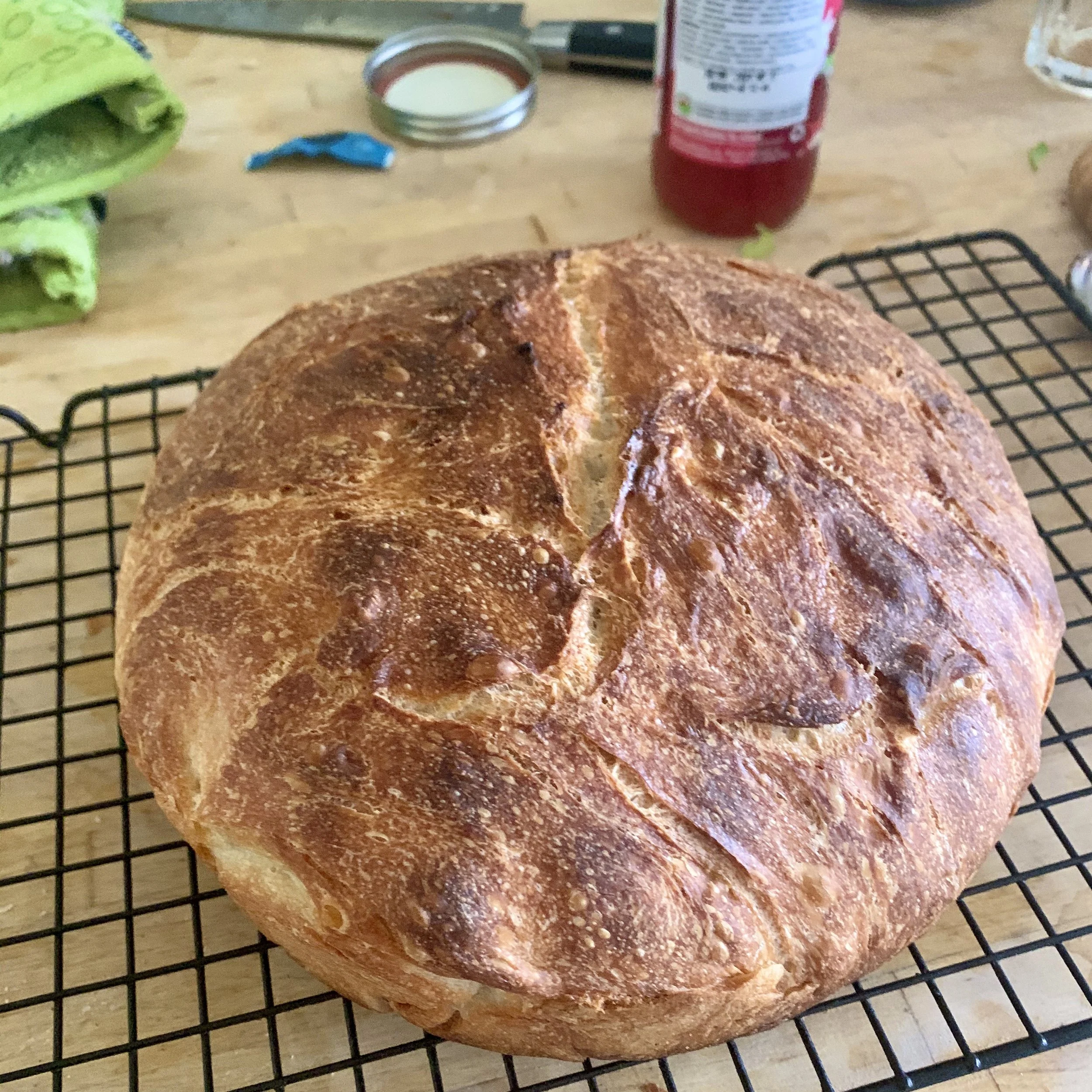 Round, freshly baked sourdough bread cooling on a black wire rack on a wooden table, with a green cloth, a can of sweetened condensed milk, a bottle of ketchup, and other kitchen items in the background.