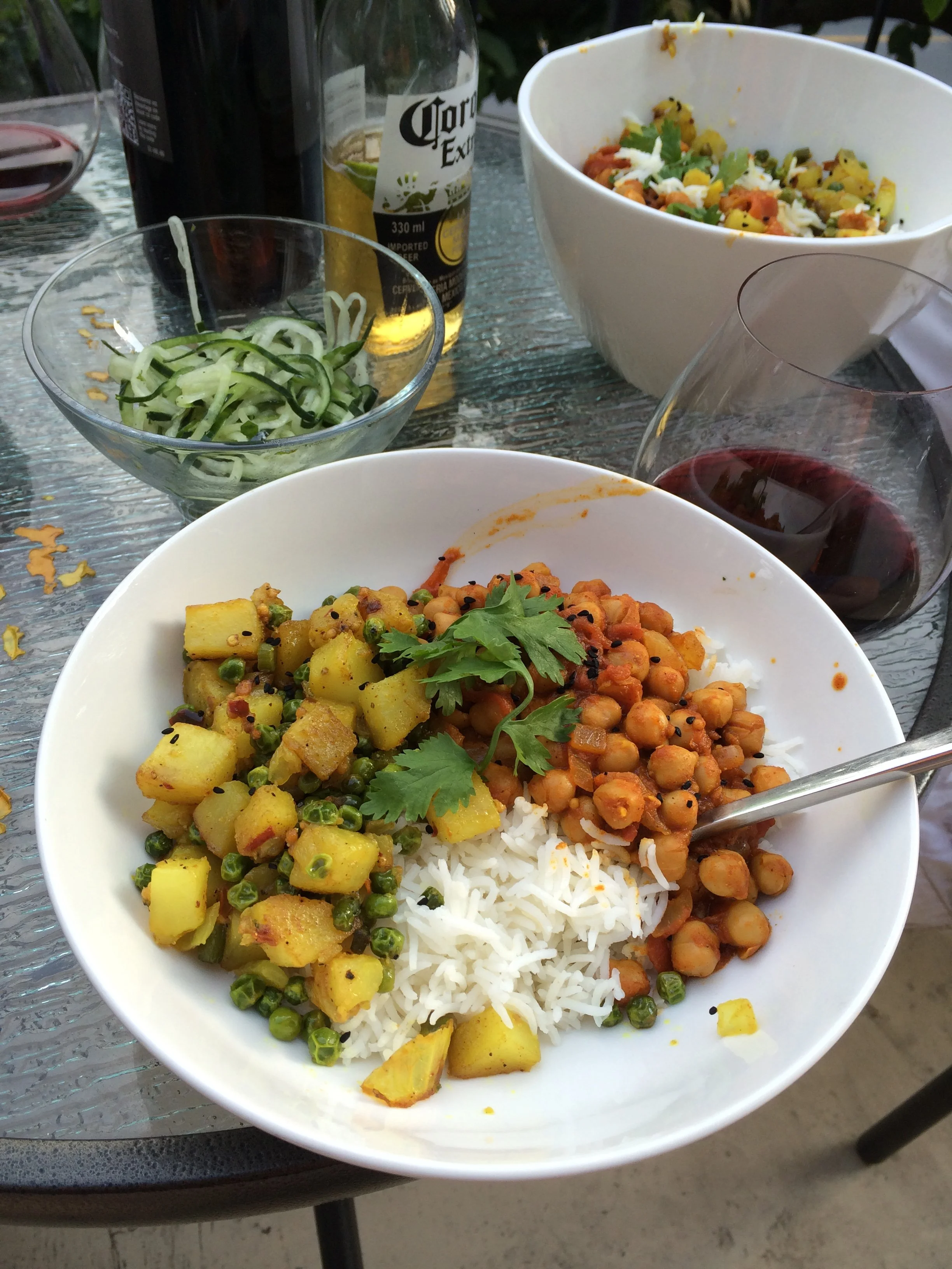 A white bowl of Indian-style vegetarian meal with cauliflower, chickpeas, rice, and yellow potatoes. Surrounding the bowl are glasses of red wine, a beer, a small glass bowl of sliced cucumbers, and another bowl of salad. The table has a glass top.