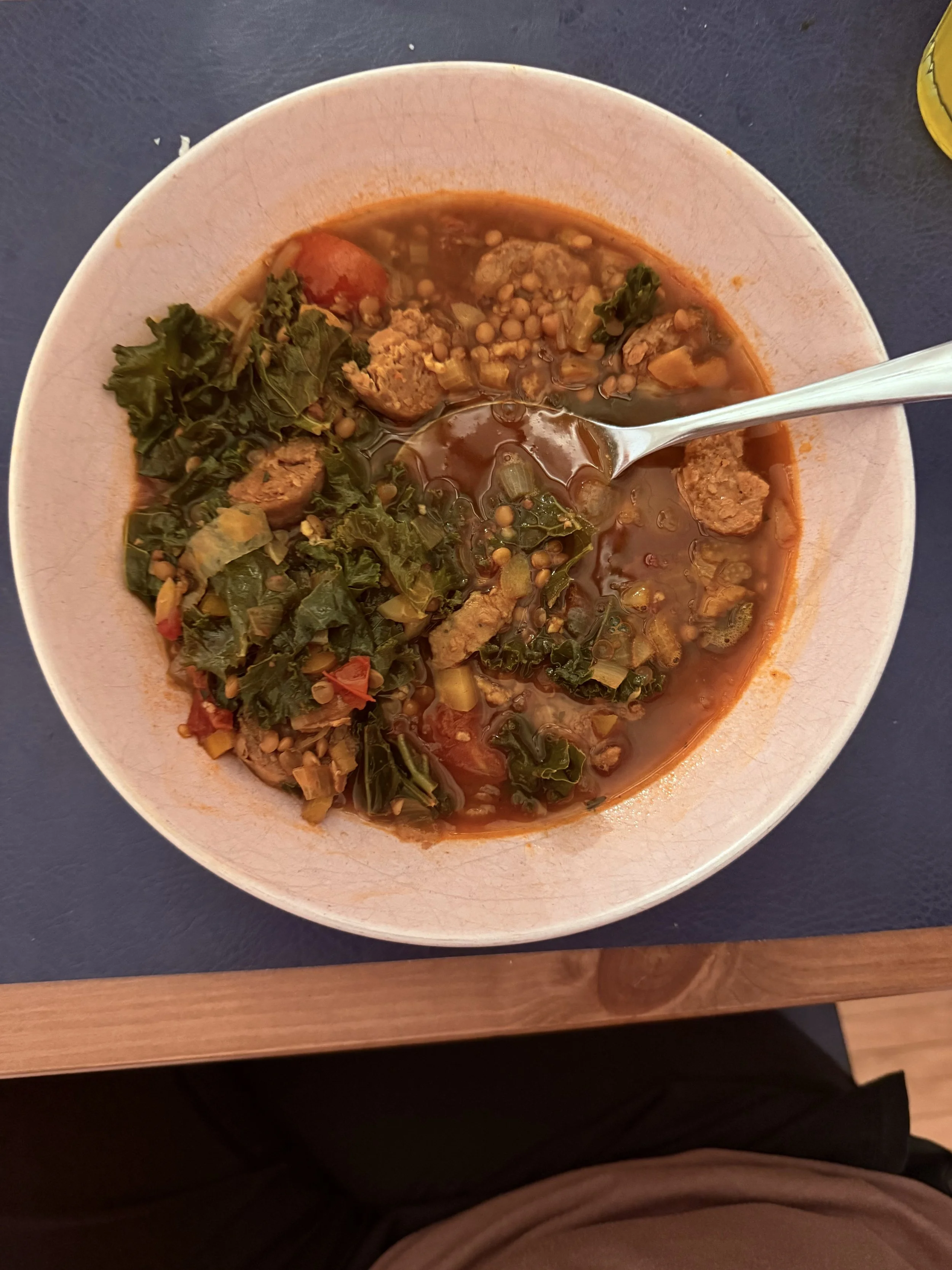 A bowl of homemade lentil soup with vegetables and greens on a blue table.