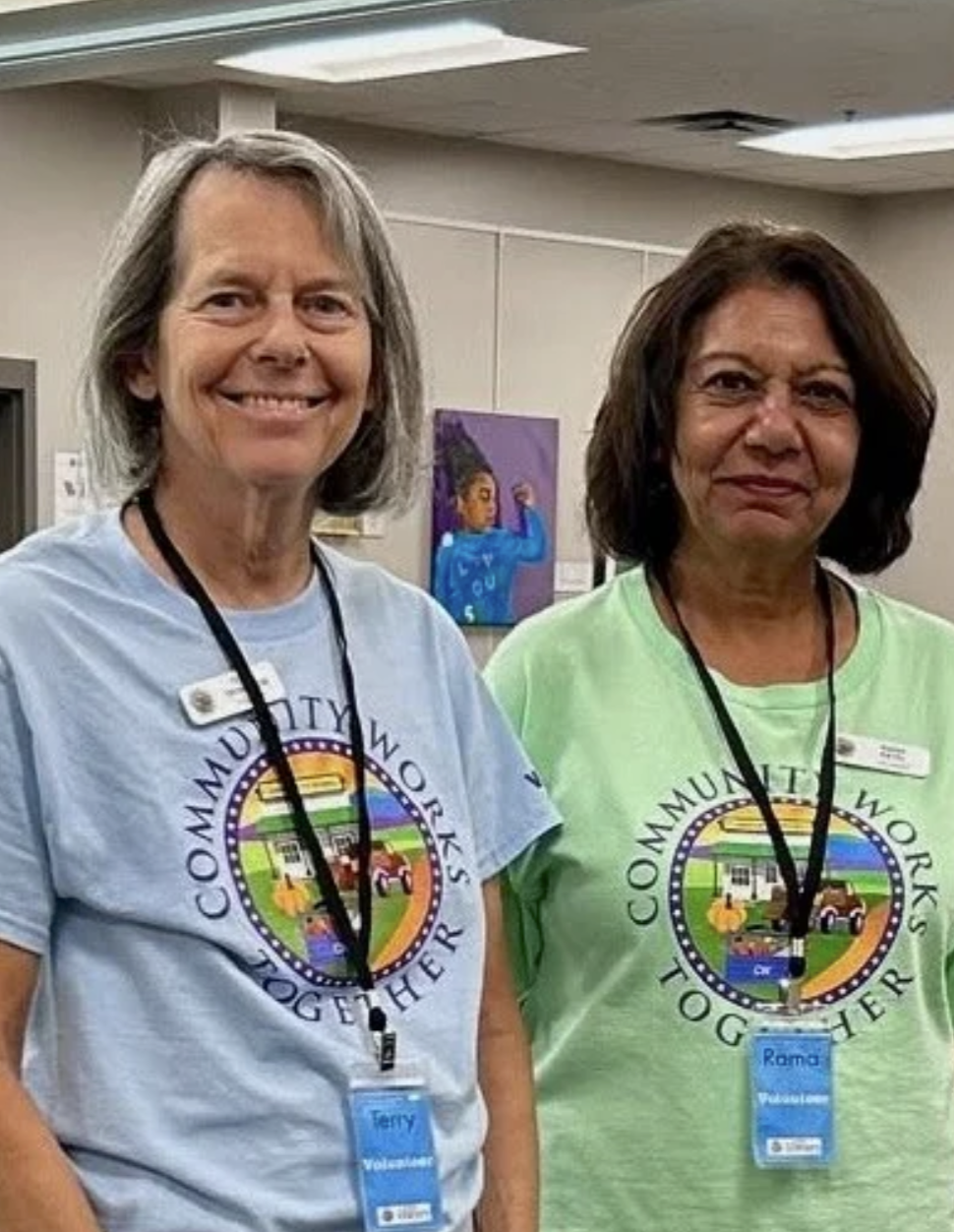 Two women wearing t-shirts with a 'Community Works Together' logo, smiling and standing side by side indoors, with artwork on the wall behind them.