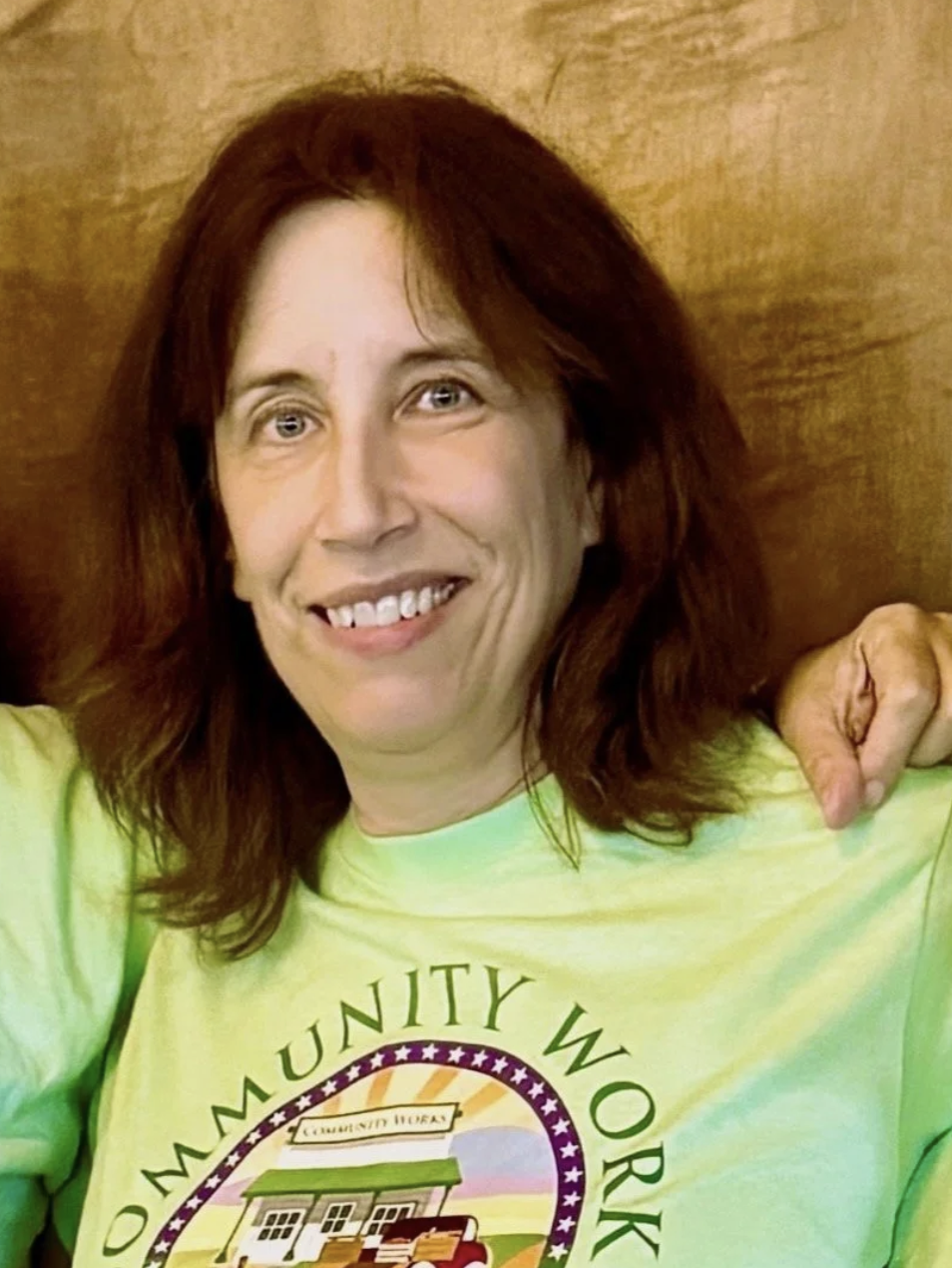 A smiling woman with shoulder-length red hair wearing a bright yellow shirt with a logo that says 'Community Work' and a graphic of a community center and a fire truck, sitting against a wooden background.