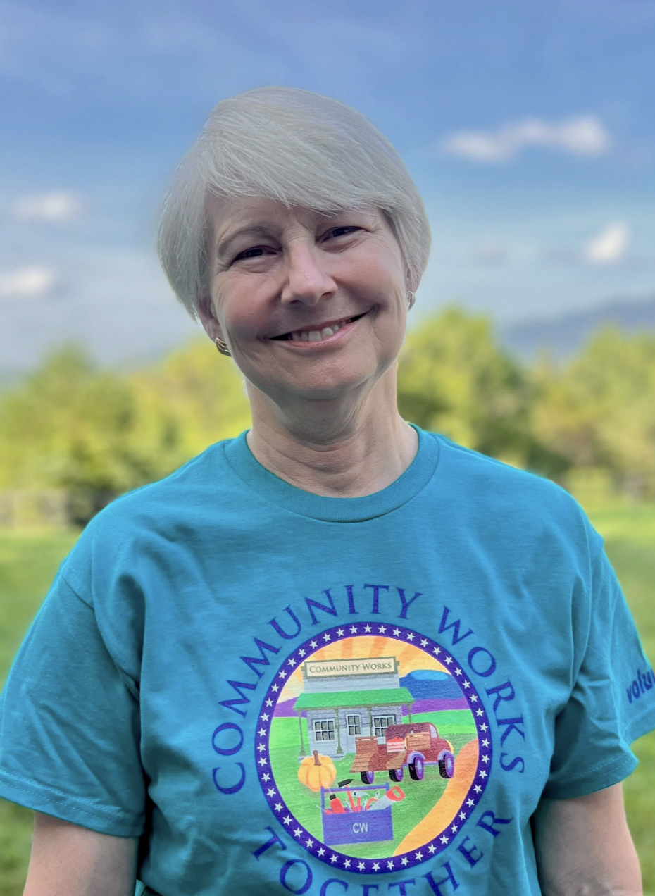 Smiling woman with short gray hair wearing a blue T-shirt with a colorful logo representing community works, standing outdoors with trees and a blue sky in the background.