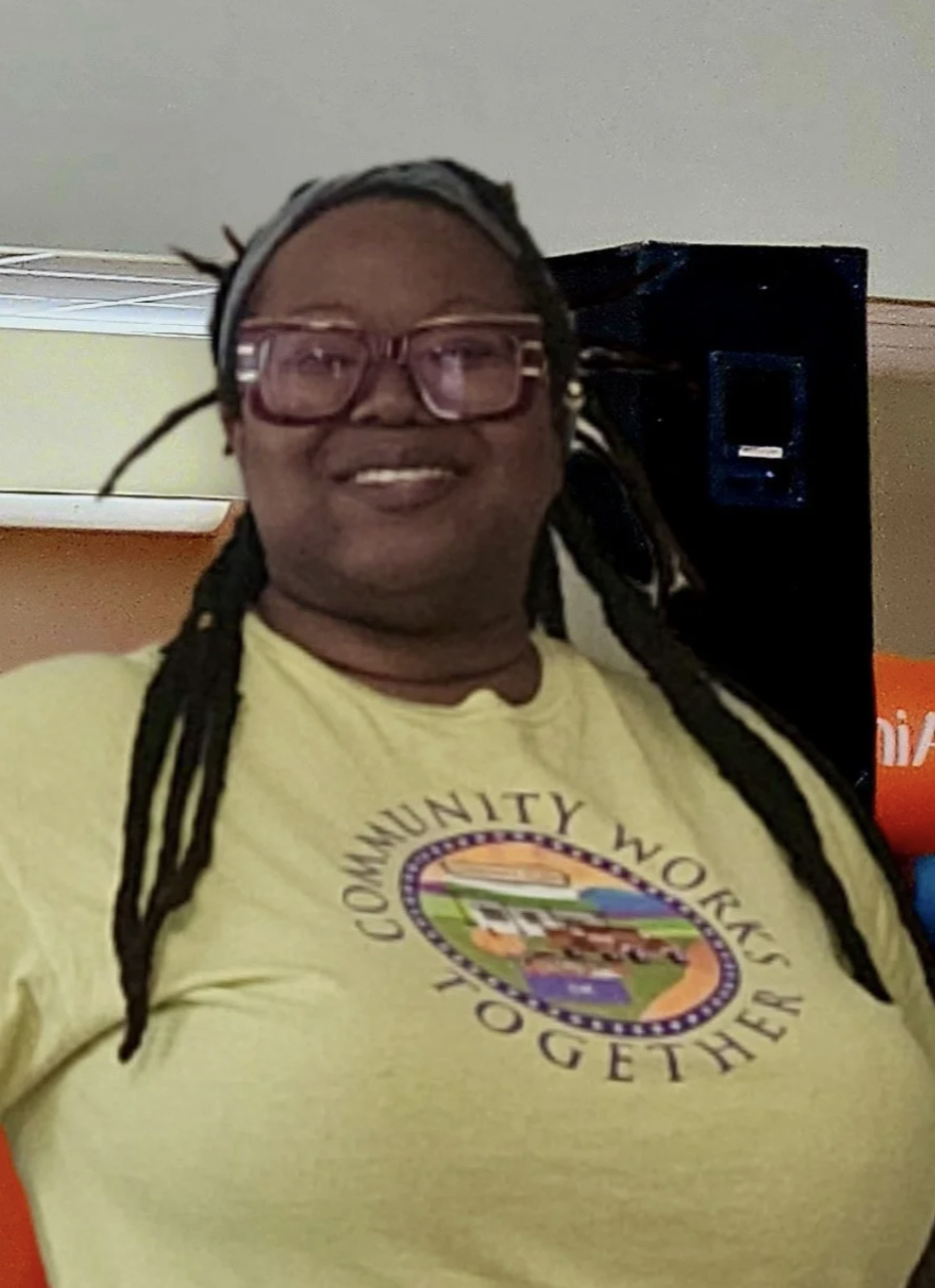 A smiling person with dreadlocks wearing glasses and a yellow T-shirt that says 'Community Works Together.' They are indoors, standing near a black speaker.