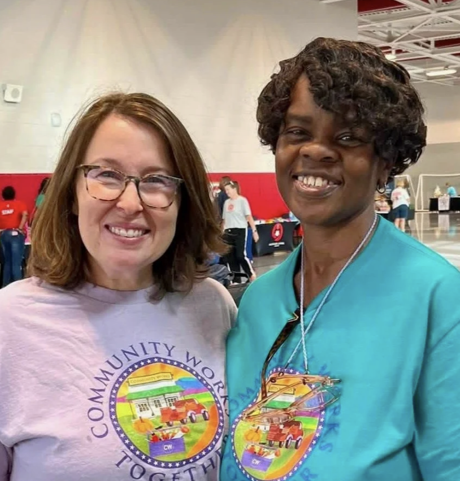 Two women smiling, standing inside a large indoor space. The woman on the left has shoulder-length brown hair and wears glasses and a light-colored t-shirt. The woman on the right has short, curly dark hair and wears a turquoise shirt with a logo. Both women are wearing large badges with colorful images and text. The background shows people and booths, suggesting a community event or gathering.