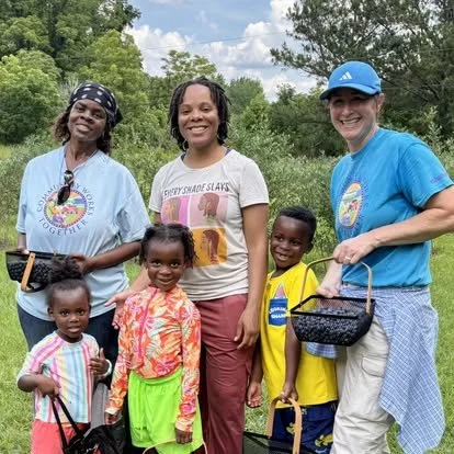 Three women and three children standing outdoors on a sunny day with trees in the background. The women are smiling, and the children are holding bags. One woman is wearing a blue t-shirt and a headband, another in a gray t-shirt with pink lettering, and the third in a blue t-shirt and a cap. The children are dressed in colorful clothes, smiling.