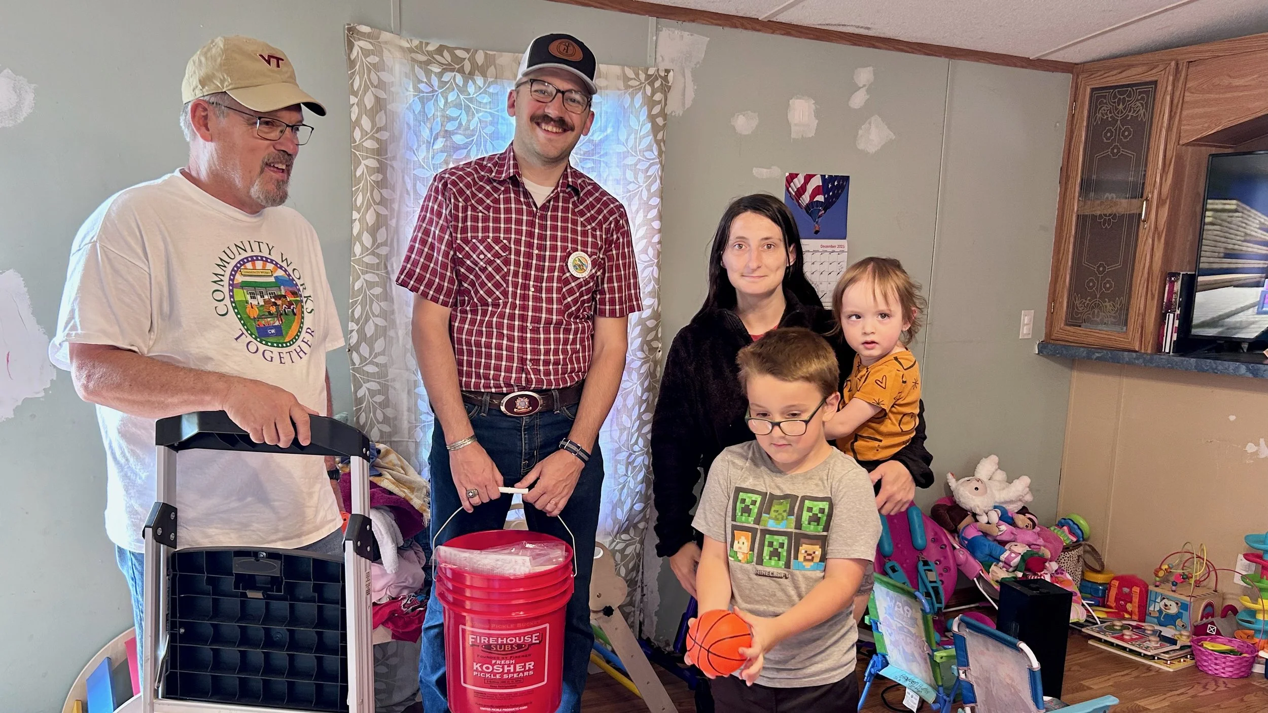 A group of five people, including two adults and three children, standing in a room filled with toys and decorations. One adult is holding a small black object, and another adult has a red bucket labeled "Firehouse Subs" and "Kosher Pickle Spear". The children are playing with toys, one holding a basketball. The room has a light green wall with patches, a patterned curtain, and a shelf filled with toys.