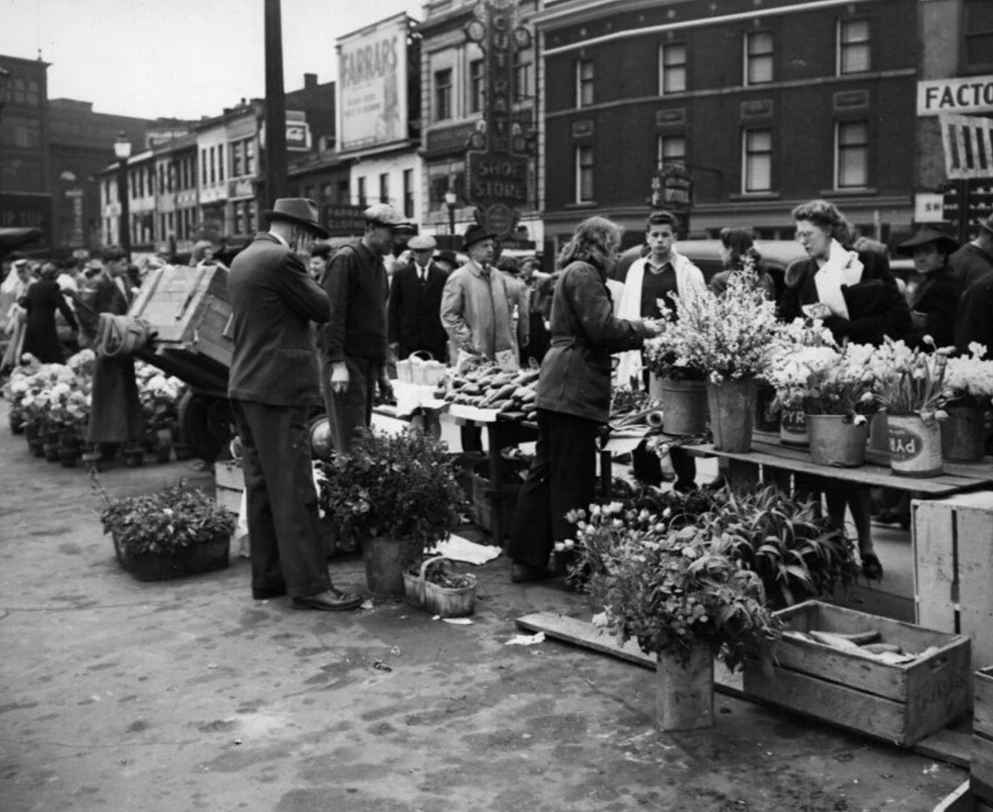 Black and white photo of a bustling city street market with people buying and selling flowers and produce, with buildings and advertisements in the background.