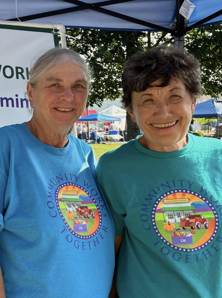 Two smiling women wearing matching turquoise T-shirts with a community work logo, standing outdoors at a festival or fair under a canopy with tents and people in the background.