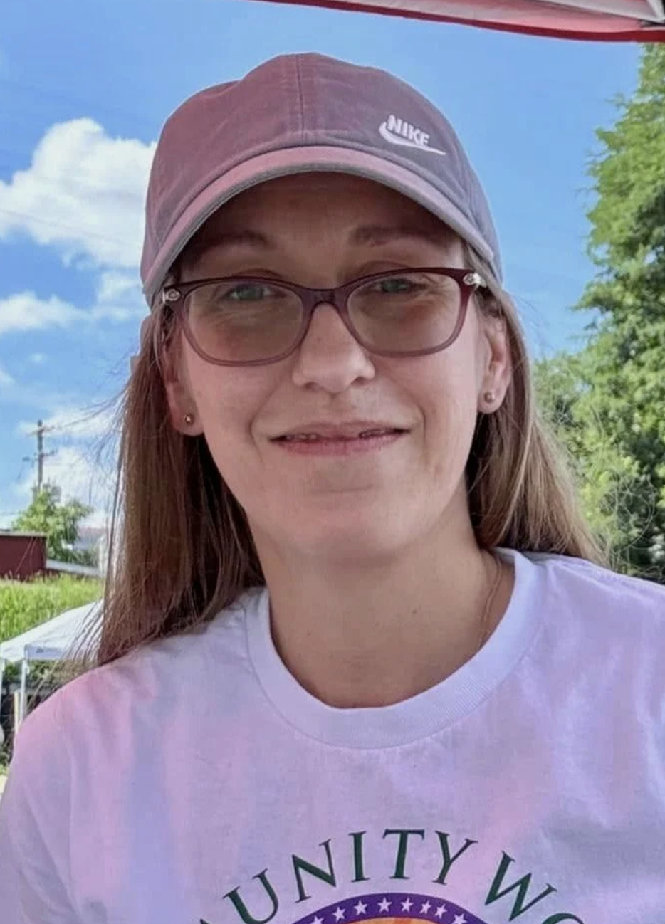 A young woman with long hair wearing a pink Nike cap, glasses, small earrings, a white T-shirt, and smiling outdoors with a blue sky, green trees, and some structures in the background.