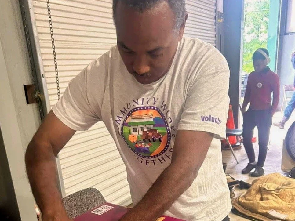 A man in a beige t-shirt with a community work logo is opening a package at a table. Two women are visible in the background near a garage door, one walking outside and another standing inside.