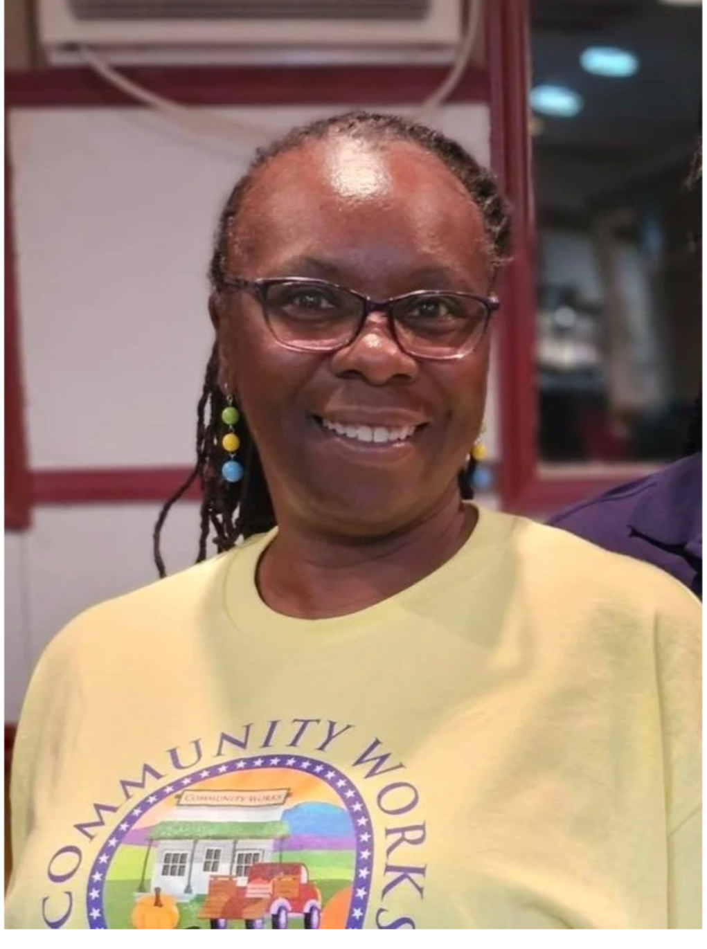 Smiling woman with glasses, earrings, and dreadlocks, wearing a yellow t-shirt with a community work logo, indoors with a red and white background.