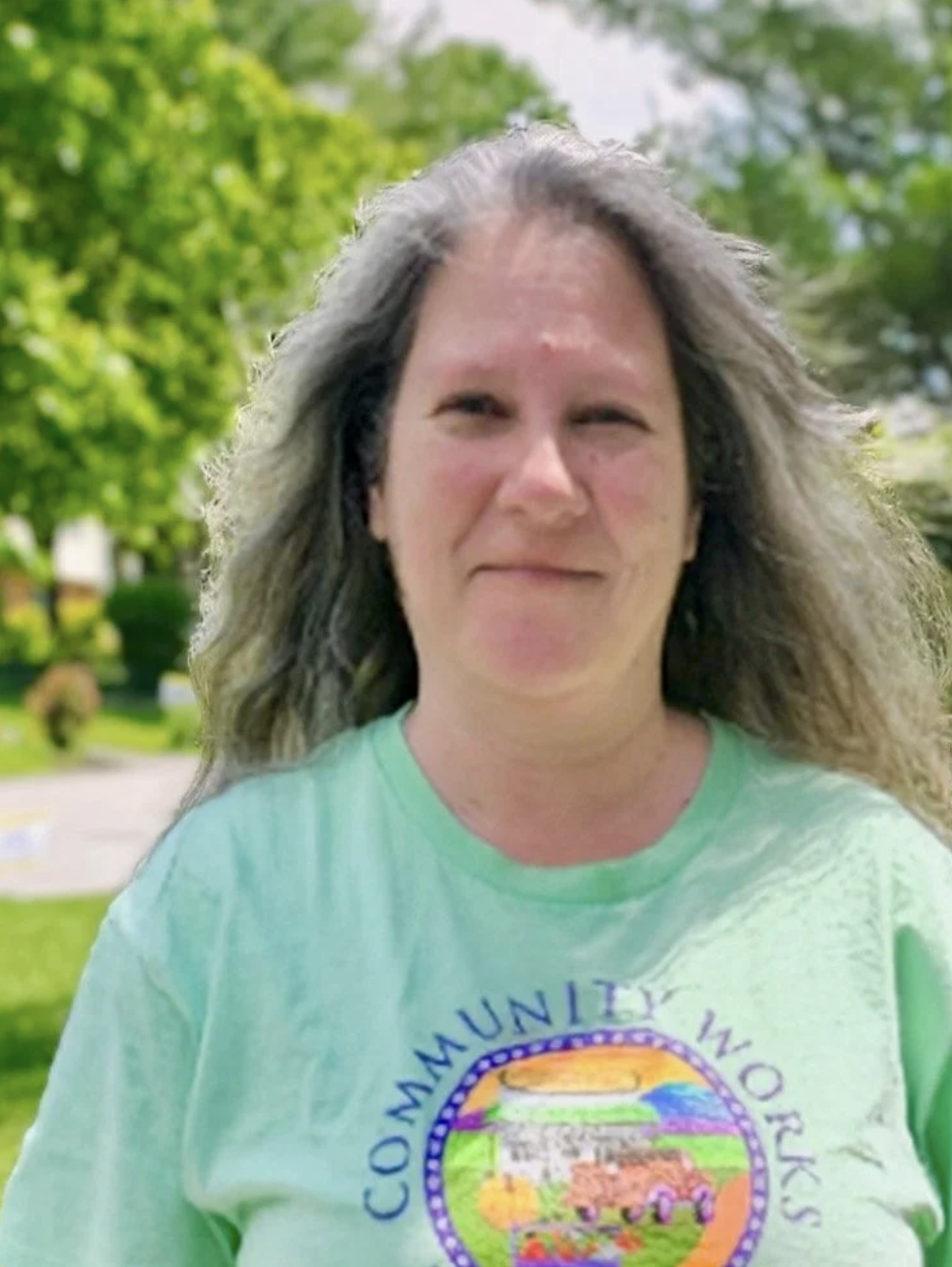 A woman with long, curly gray hair outdoors, wearing a light green T-shirt with a colorful community logo. Green trees and bushes are in the background.