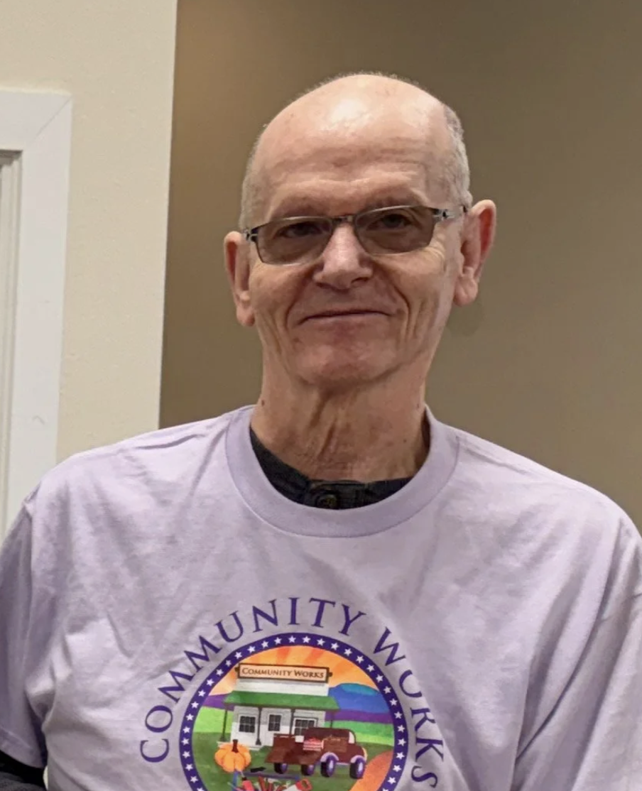 An elderly man wearing glasses and a light purple T-shirt with a colorful logo that reads "Community Wucks", featuring a barn, tractor, and farm scene, standing indoors against a beige wall.
