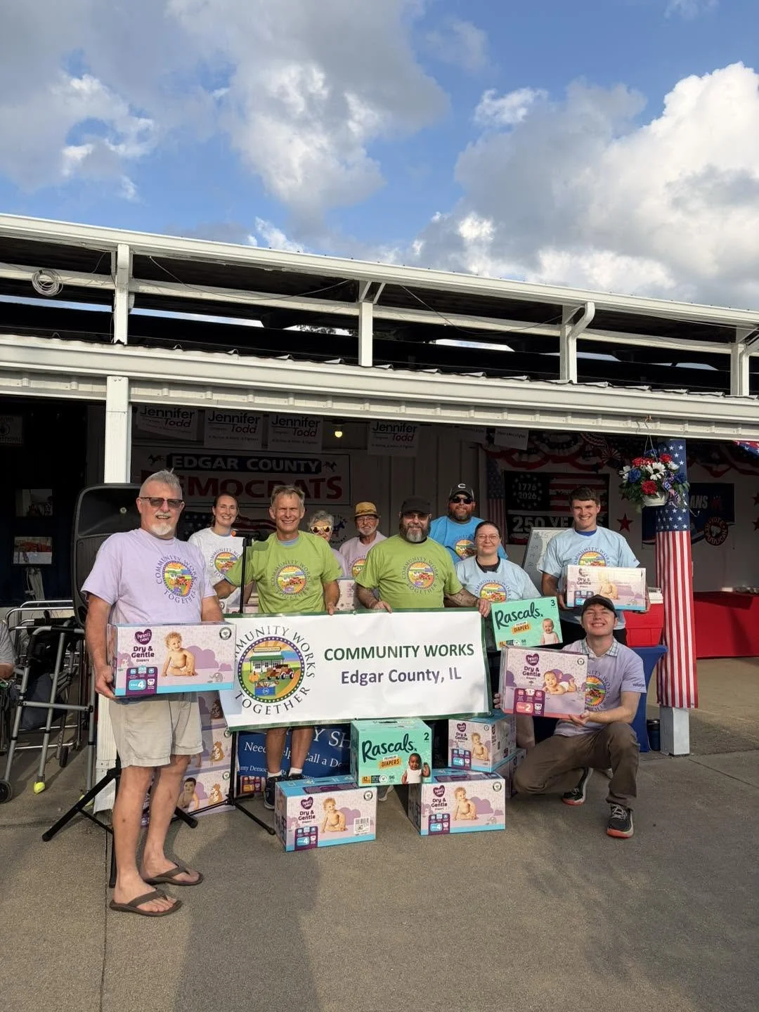 Group of people holding a community works banner with diaper boxes in front, standing in front of a stage with red, white, and blue decorations, at a patriotic event in Edgar County, Illinois.