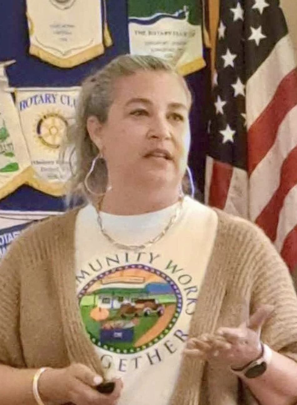 A woman speaking, wearing a white T-shirt with a colorful logo and text that says 'Community, World, Together.' She has gray hair styled back, large hoop earrings, a gold chain necklace, and a brown sweater. Behind her are American flags and several banners, indicating an event or meeting setting.