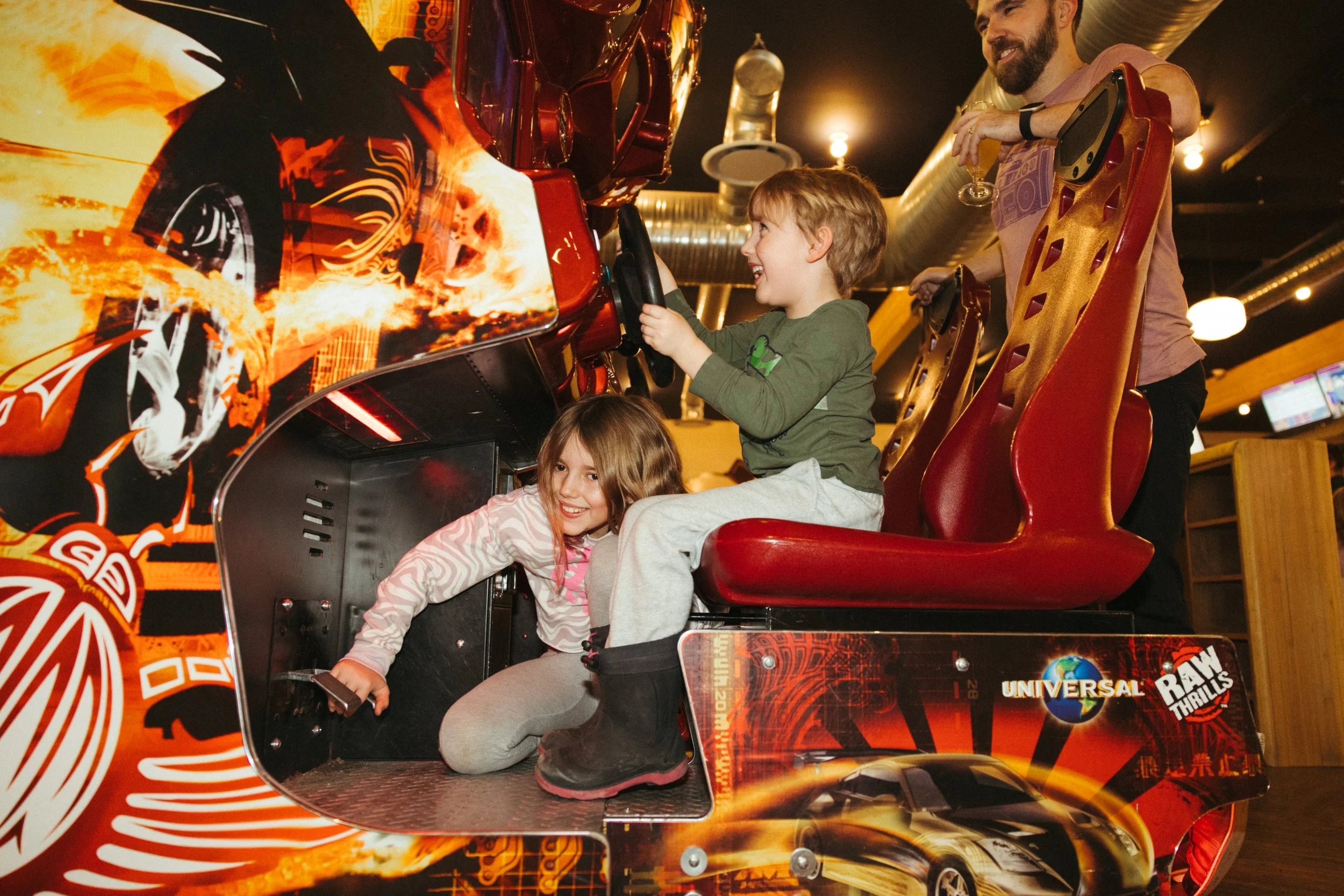 Two children riding inside a virtual reality racing simulator at an arcade, with a man standing behind them. The video game scene features a high-tech race car and fiery visuals, with the arcade's branding and logo visible.