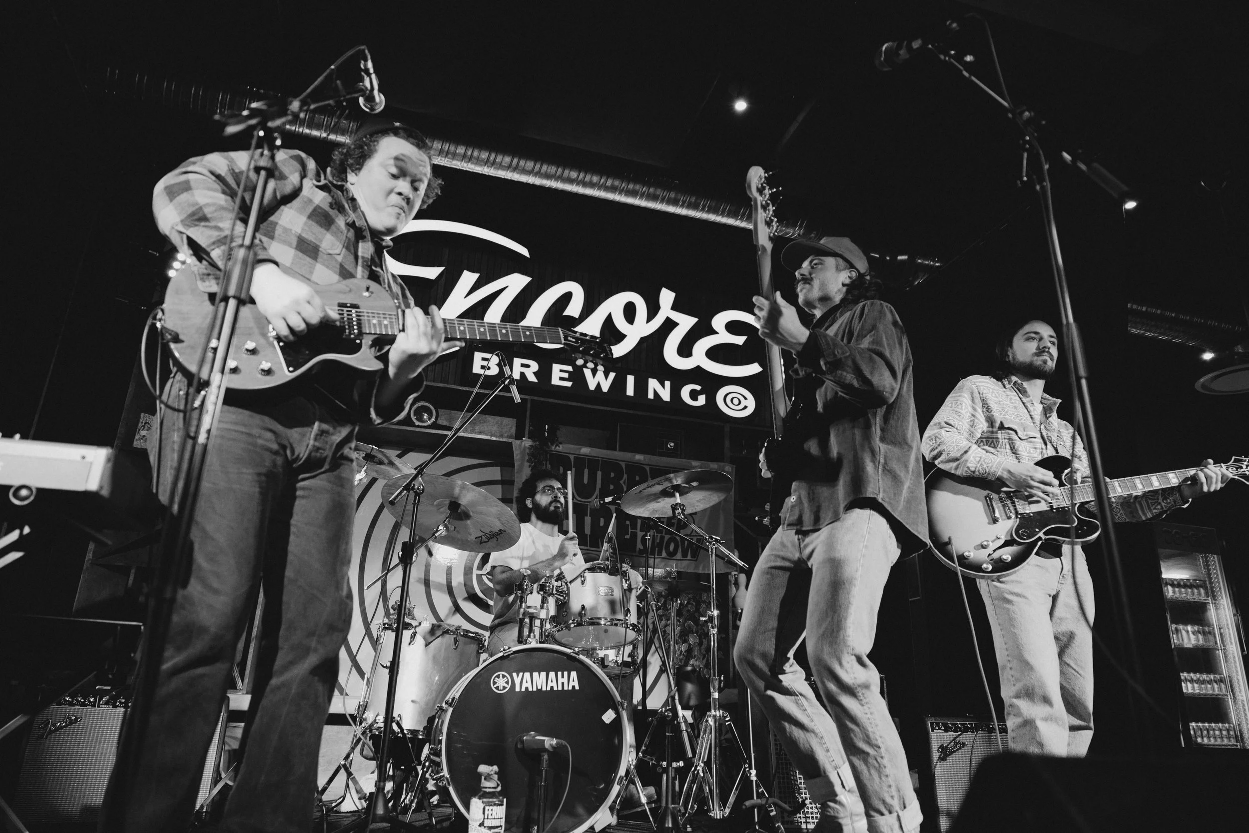 A band performing live on stage at a bar with a sign that reads 'Encore Brewing'. The band consists of four members: a guitarist on the left, a drummer in the back, a singer with a cap and a guitar on the right, and another guitarist. The scene is in black and white.