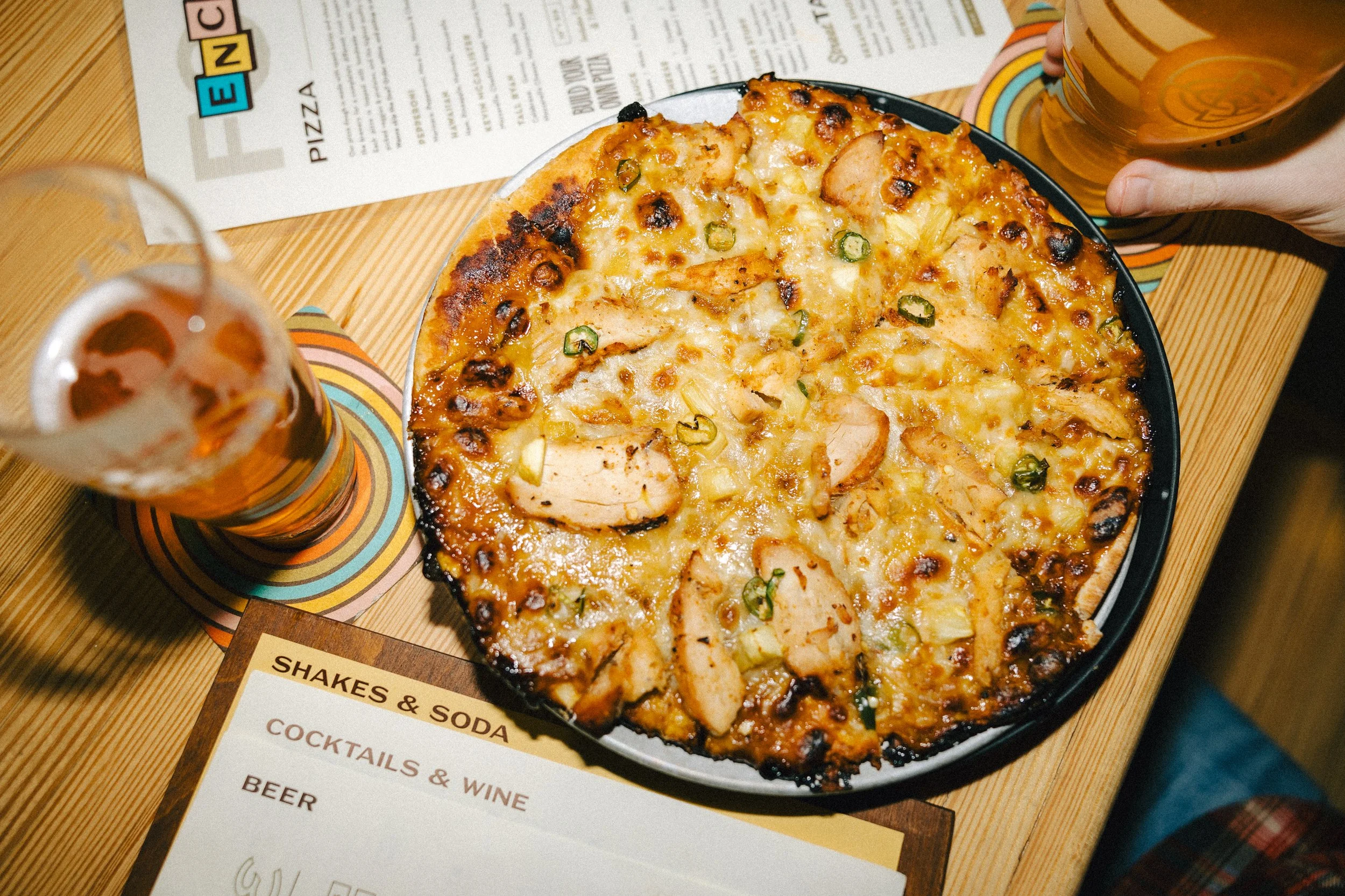 A pizza with chicken, green peppers, and cheese on a wooden table with two glasses of beer and a menu.