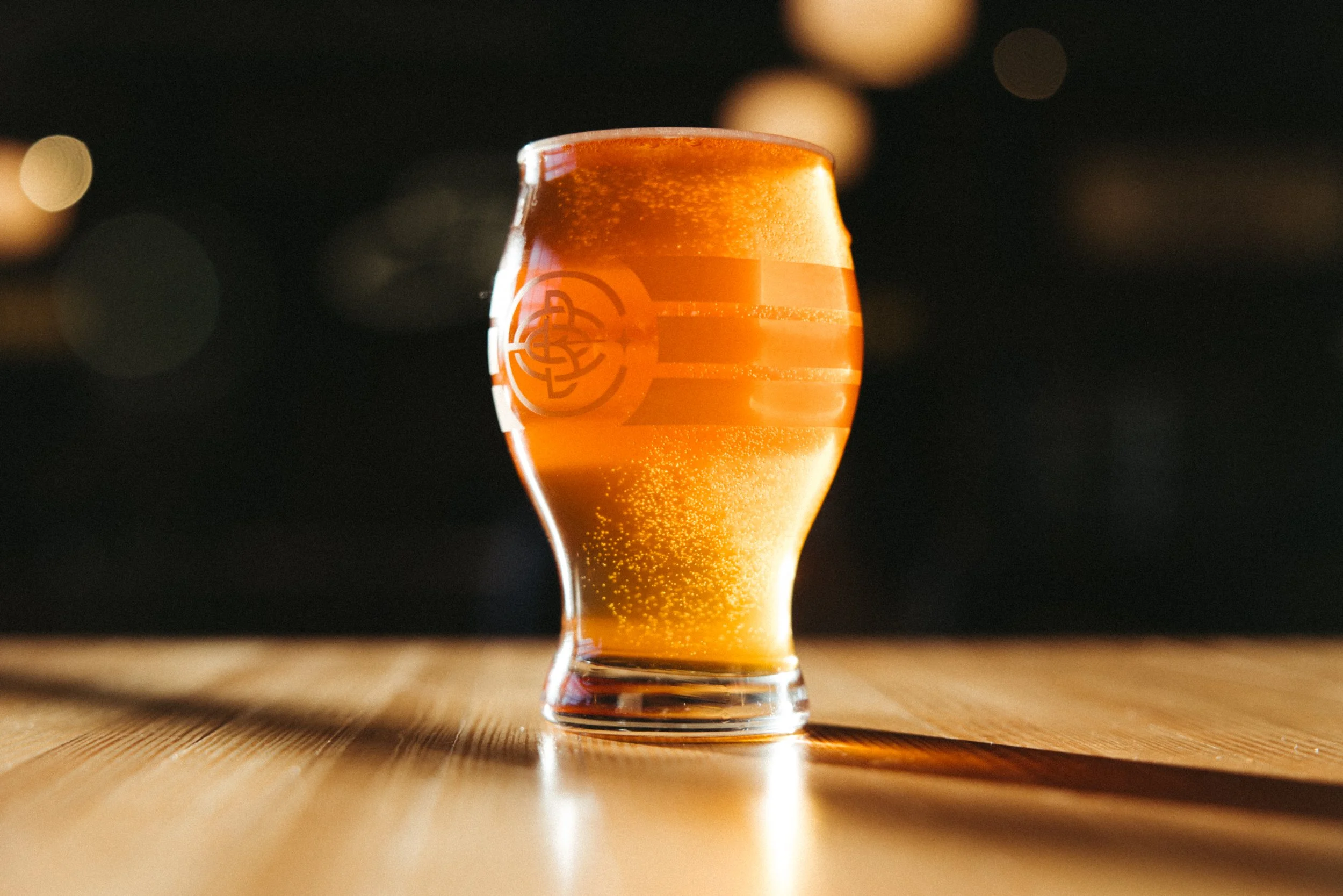 A tulip-shaped glass of beer with a logo on the side, sitting on a wooden surface with a dark, blurry background.