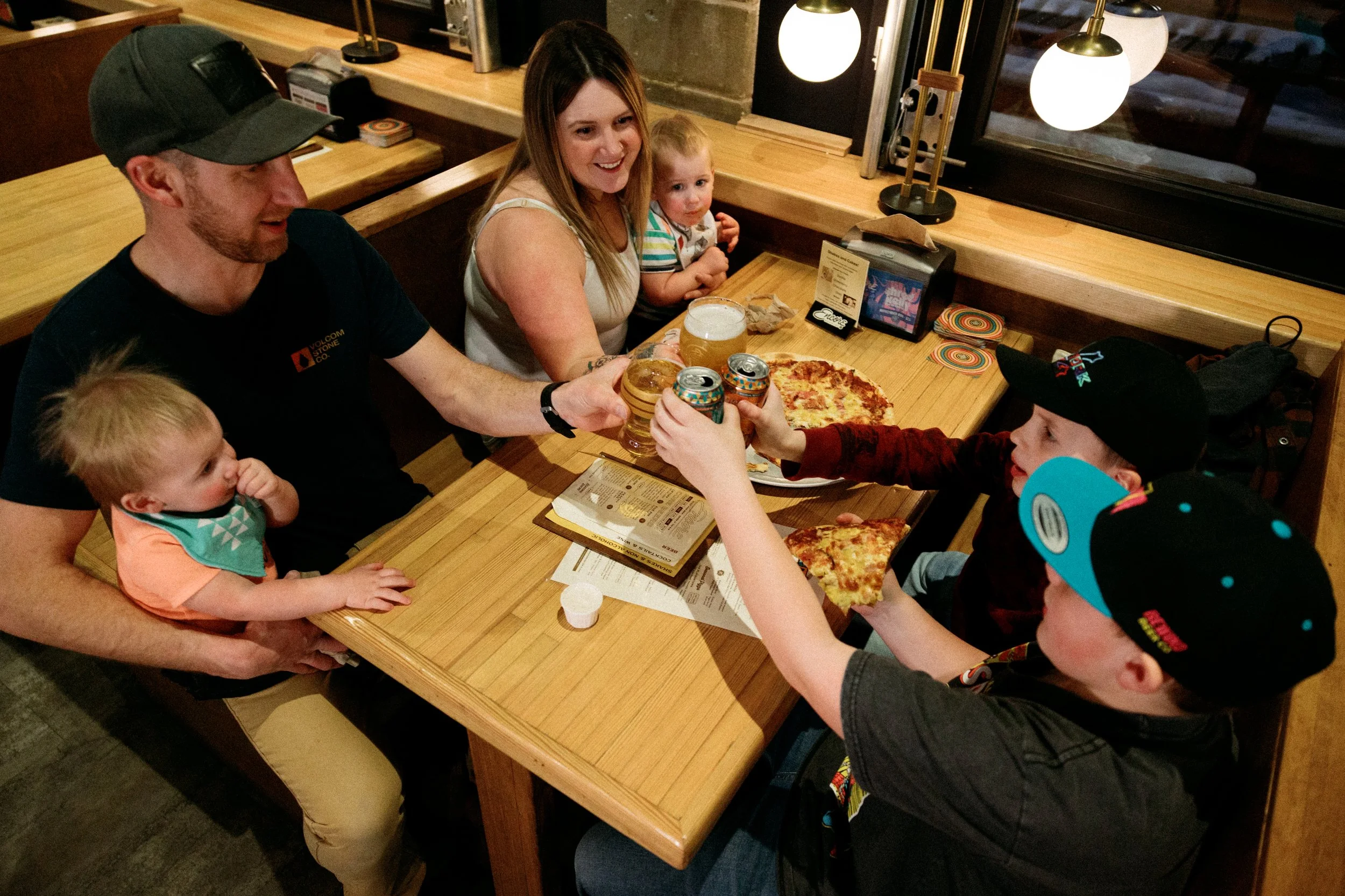 A family celebrating at a restaurant, raising drinks in a toast, with two children and a pizza on the table.