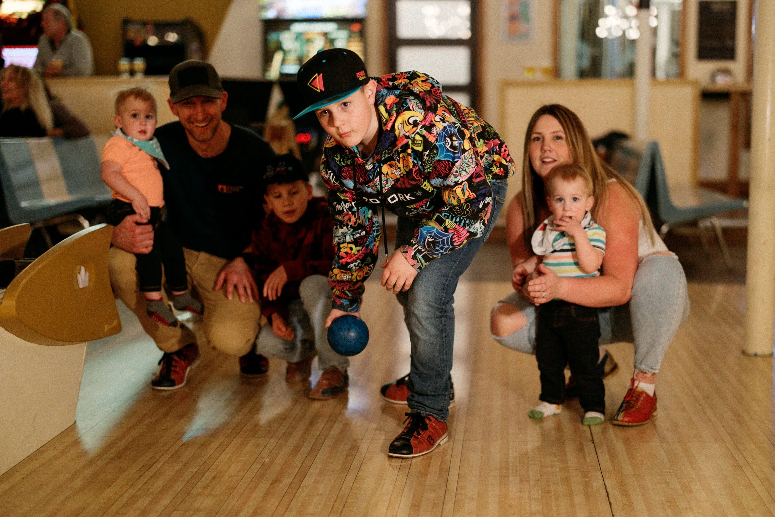 A young boy bowls at a bowling alley, with a group of children and adults watching. The boy is wearing a colorful jacket and orange shoes, holding a blue bowling ball. The group is gathered around him, with some children sitting and standing, looking at the boy.