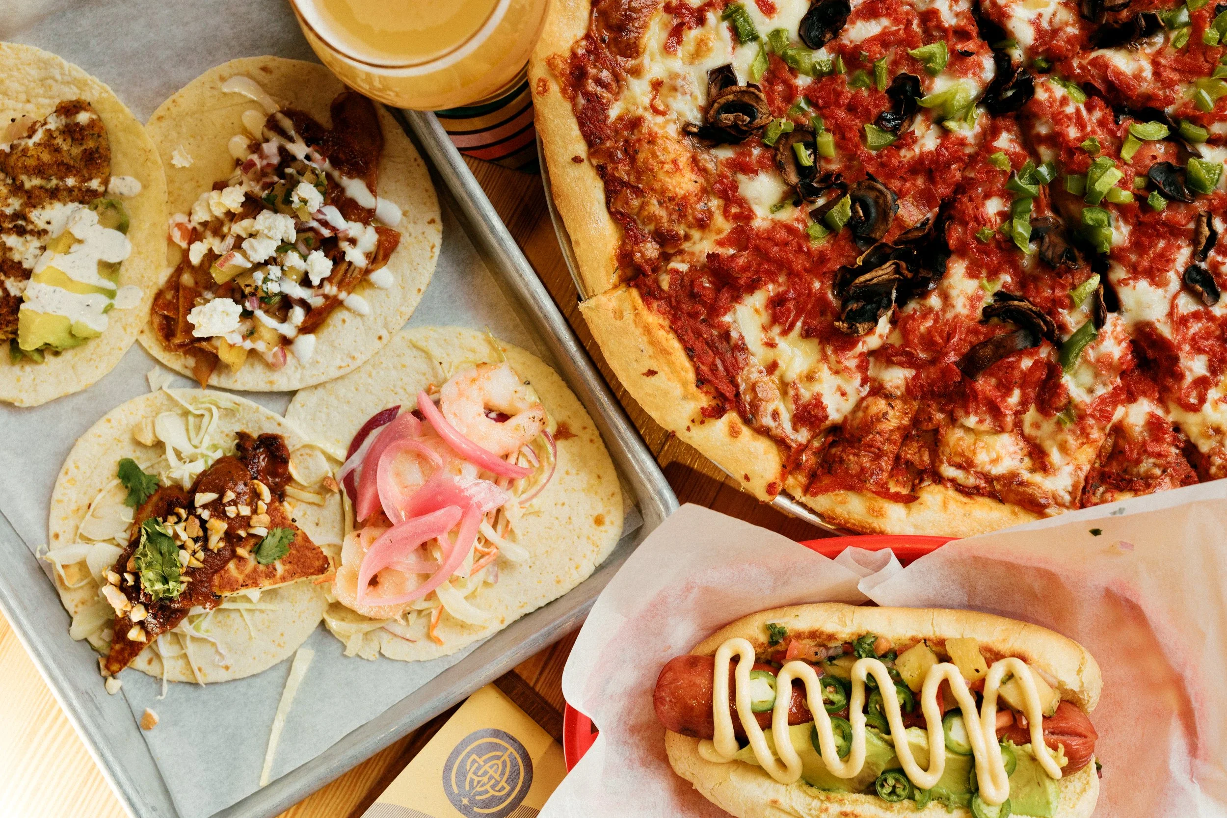 Assorted Mexican food including tacos with various toppings, a large pepperoni pizza with black olives and green peppers, and a hot dog with mustard, lettuce, tomato, and jalapenos.