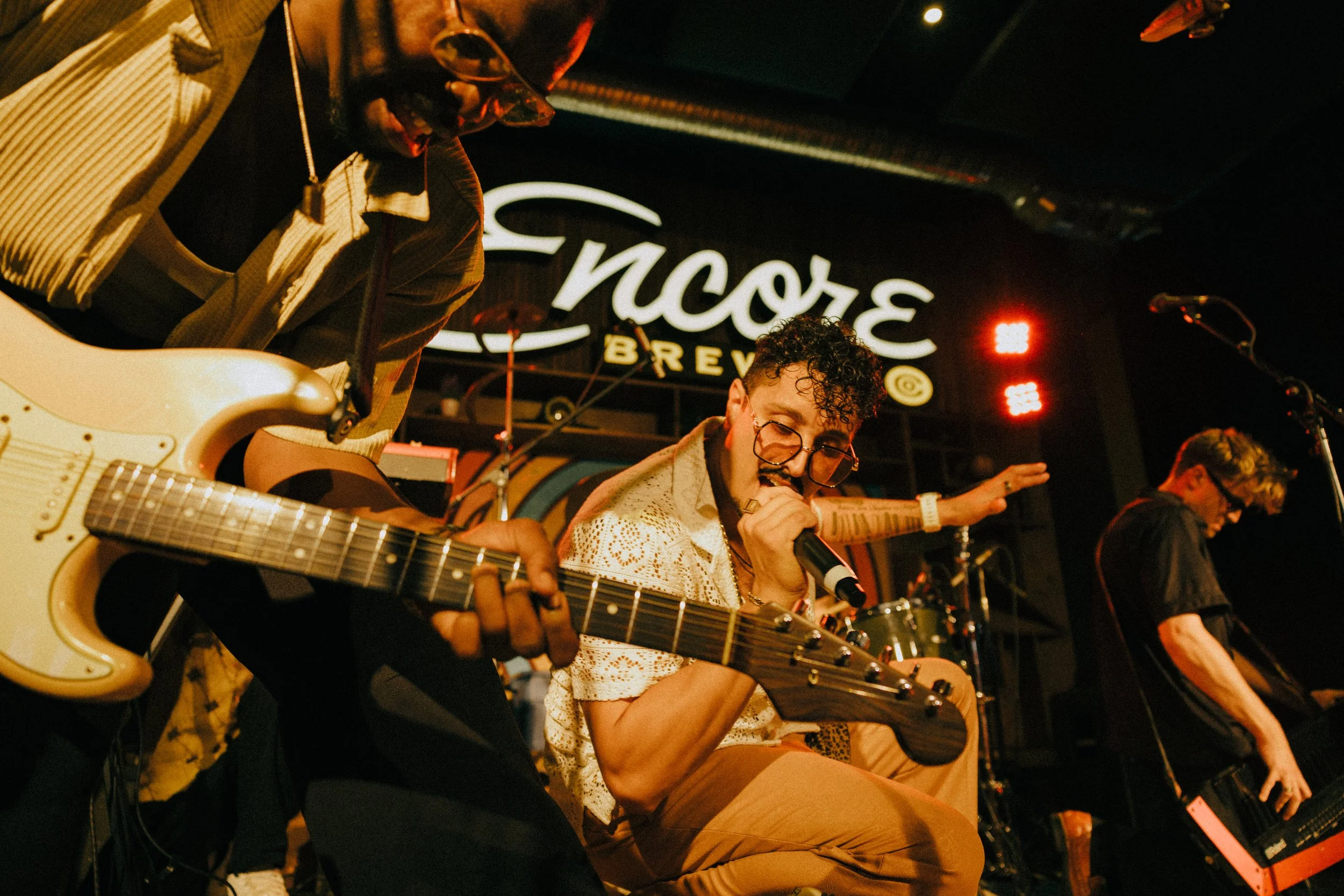 Musicians performing on stage at a venue with a sign that says 'Fino's BREW' in the background. One musician is playing an electric guitar, another is singing into a microphone, and a third is seen with a music stand. The scene is illuminated with stage lights.