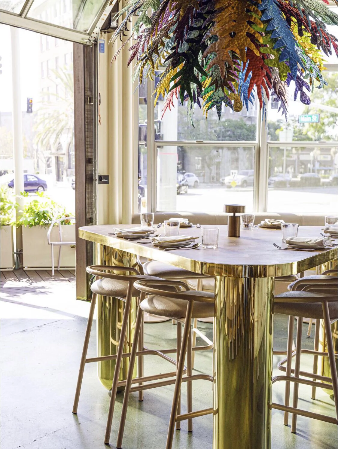 Interior of a stylish restaurant with a high-top table with a gold metallic base, set with plates, glasses, and napkins. There are four beige bar stools around the table. Above the table, a colorful, leafy pendant light hangs. Large windows in the ba