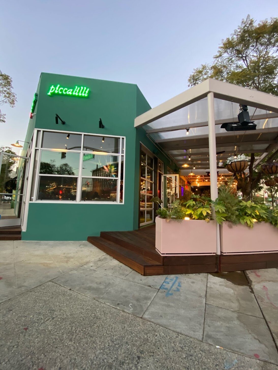 Exterior of a modern restaurant with a green facade and neon sign reading 'piccalii'. There is an outdoor patio with potted plants and hanging flower baskets, and the interior is visible through large windows.