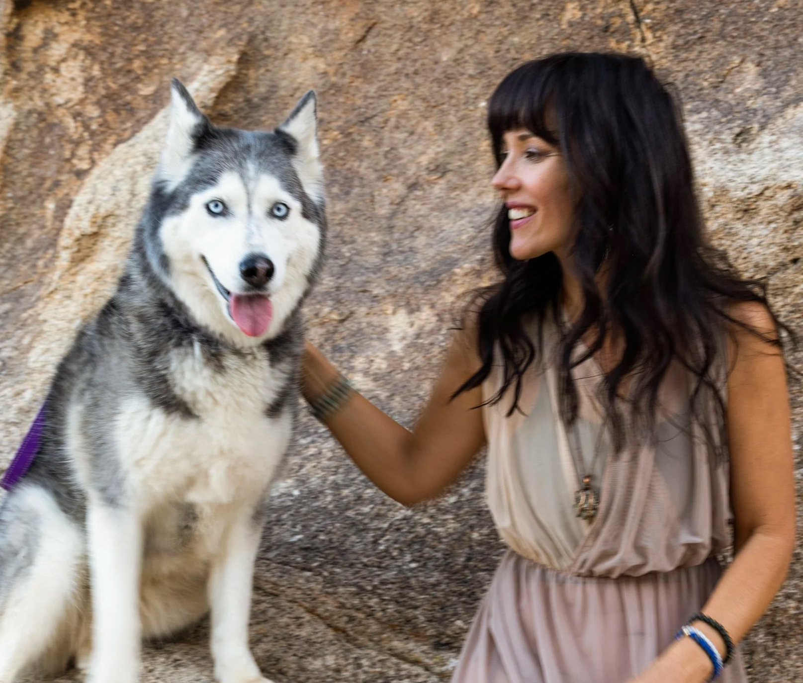A woman with dark wavy hair smiling and petting a Siberian Husky dog with blue eyes and a black-and-white coat in front of a rocky background.