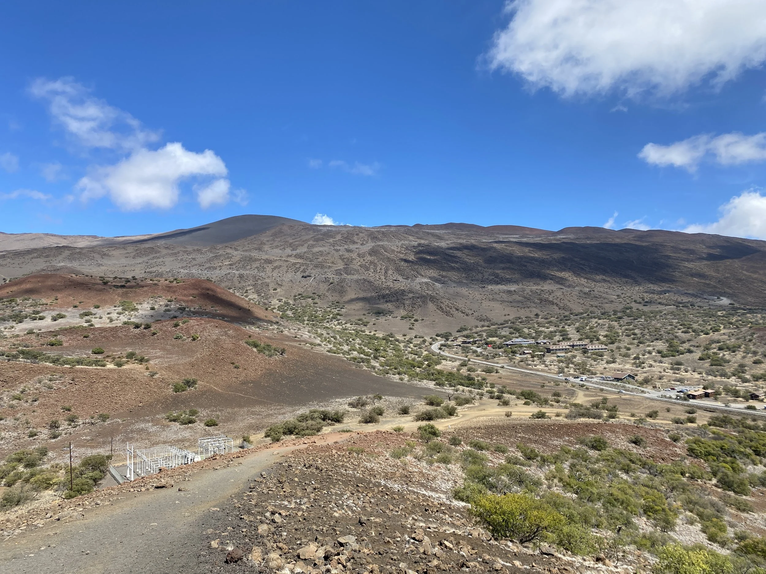 A mountainous landscape with brown and gray hills, sparse green vegetation, a clear blue sky with some scattered clouds, and a winding road with buildings and vehicles in the distance.