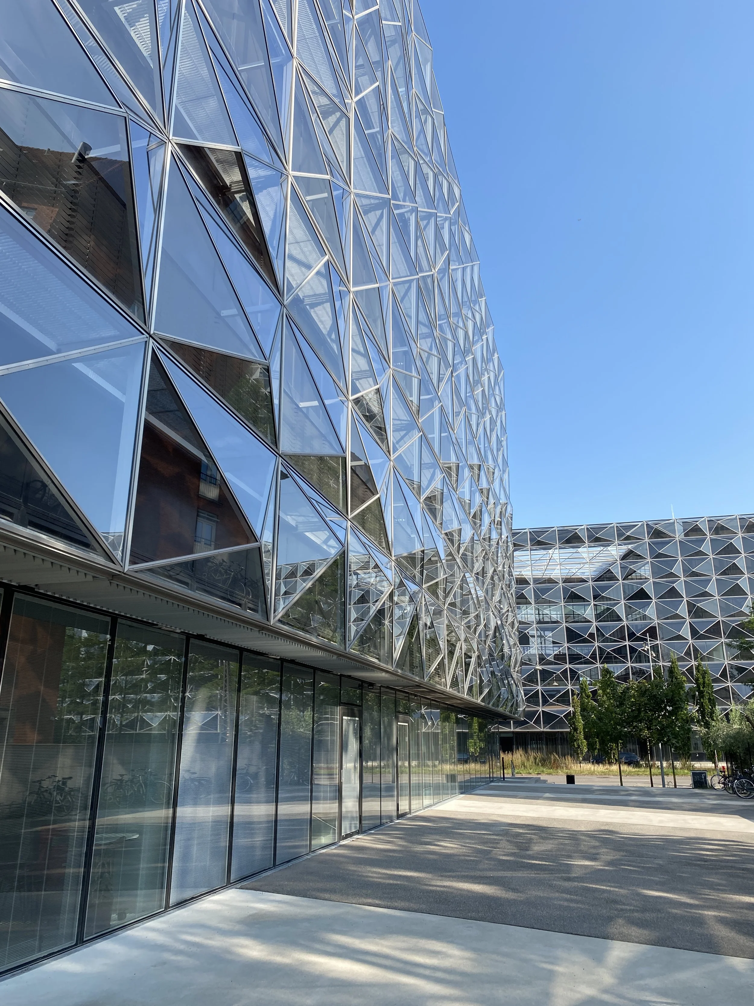 Modern building with a glass facade featuring geometric patterns, set against a clear blue sky with a paved area and trees in the background.