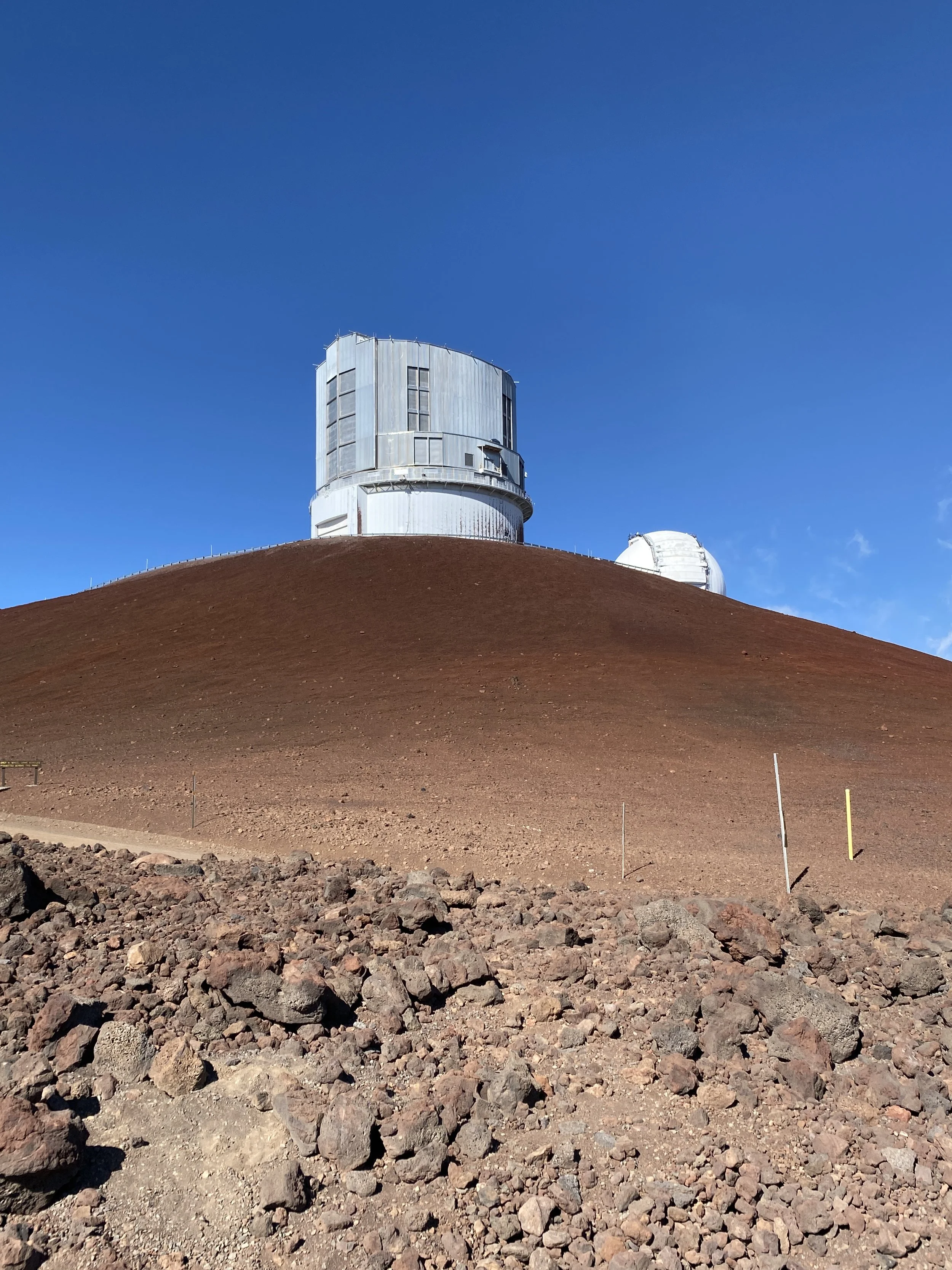 A mountain with a rocky base and reddish slope featuring an observatory with two large telescope domes on top, under a clear blue sky.