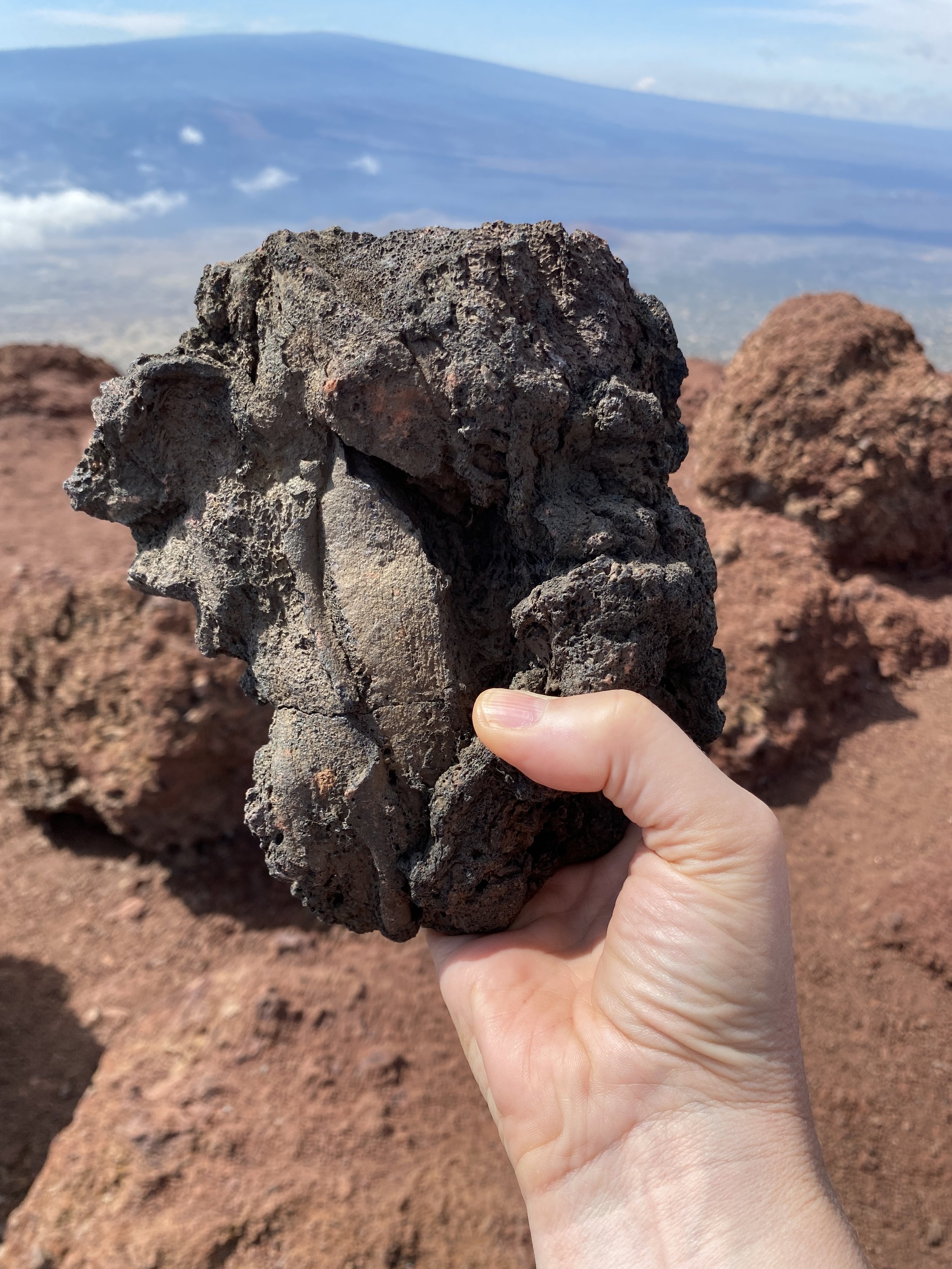 Close-up of a person's hand holding a rough, dark volcanic rock with a textured surface, against a rocky, desert-like landscape and a clear blue sky in the background.