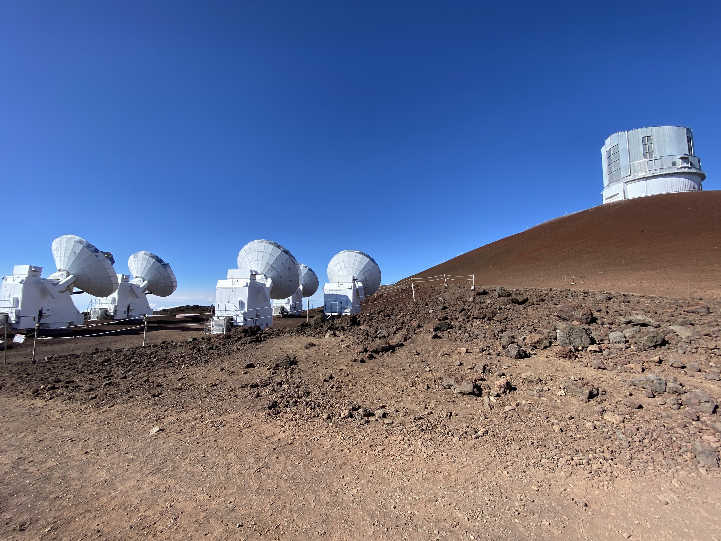 A clear blue sky over a mountain with a series of large white satellite dishes and a round observatory structure on a brown, rocky surface.