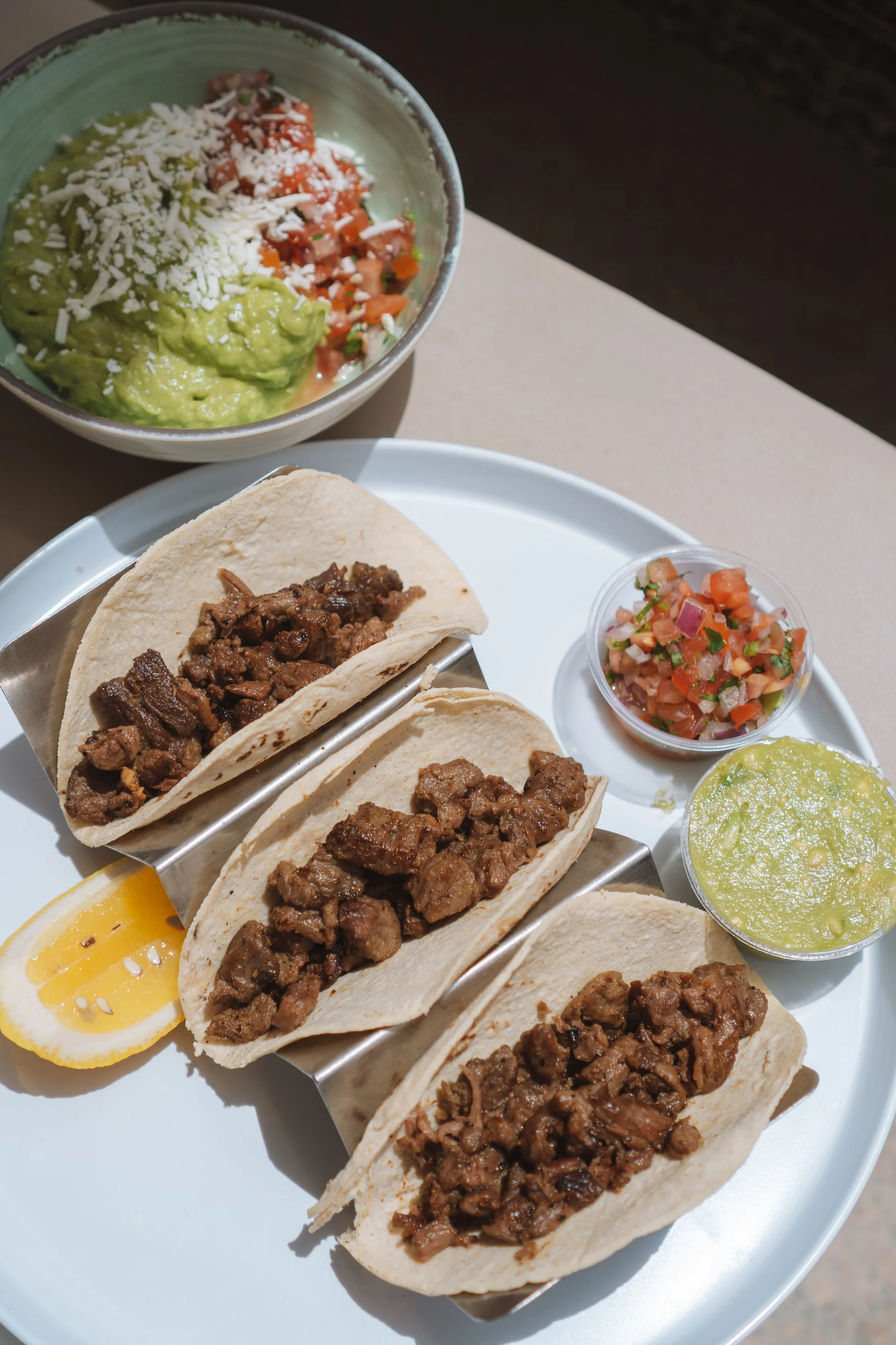A plate with four beef tacos topped with chopped meat, accompanied by a lemon wedge and green sauce. In the background, there is a bowl of guacamole with shredded cheese and a mini cup of pico de gallo.