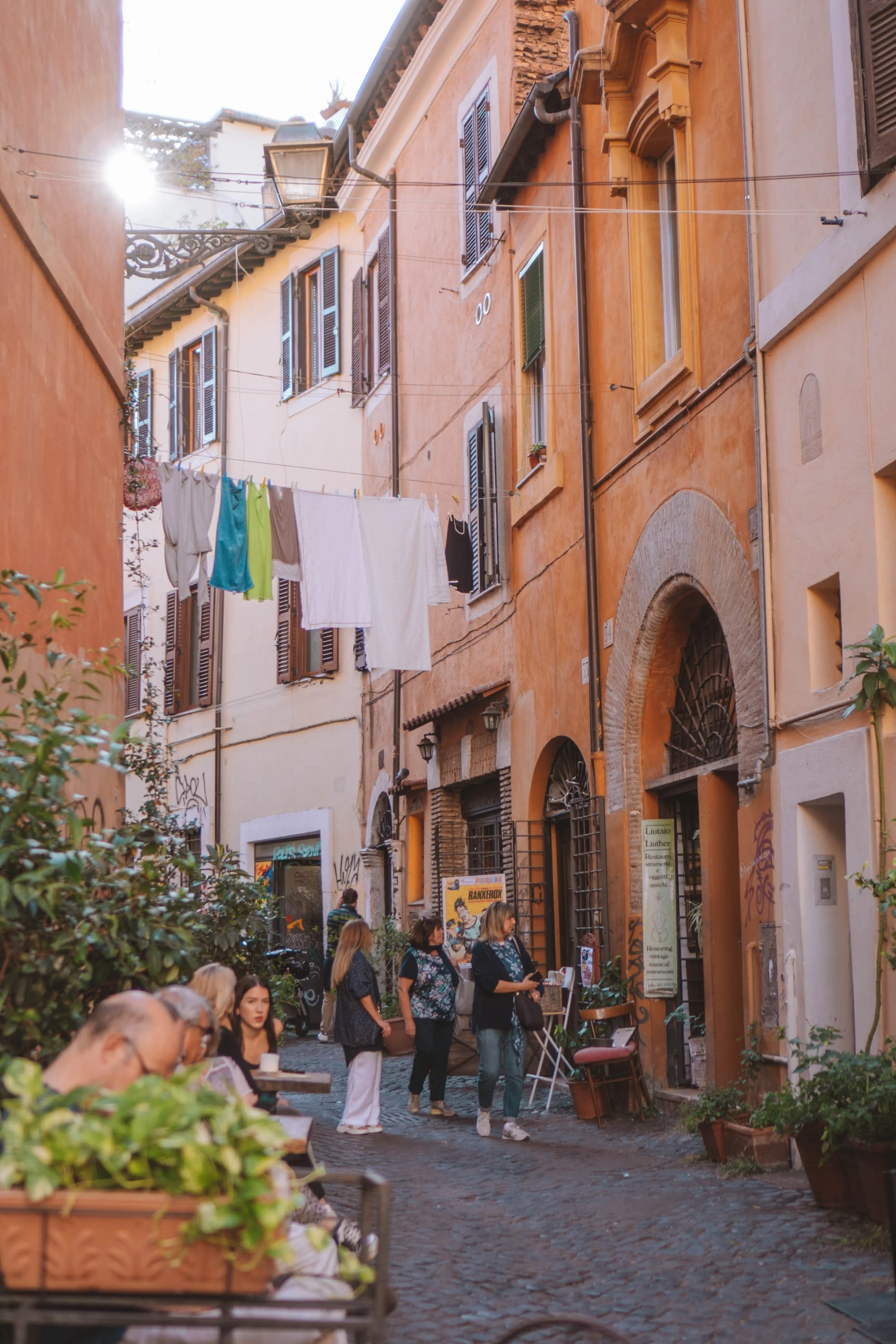 A European street scene with people walking and sitting at outdoor cafes. The buildings are painted in warm colors like ochre and terracotta, with wooden shutters on the windows. Clothes are hanging on lines strung across the street, and there are si