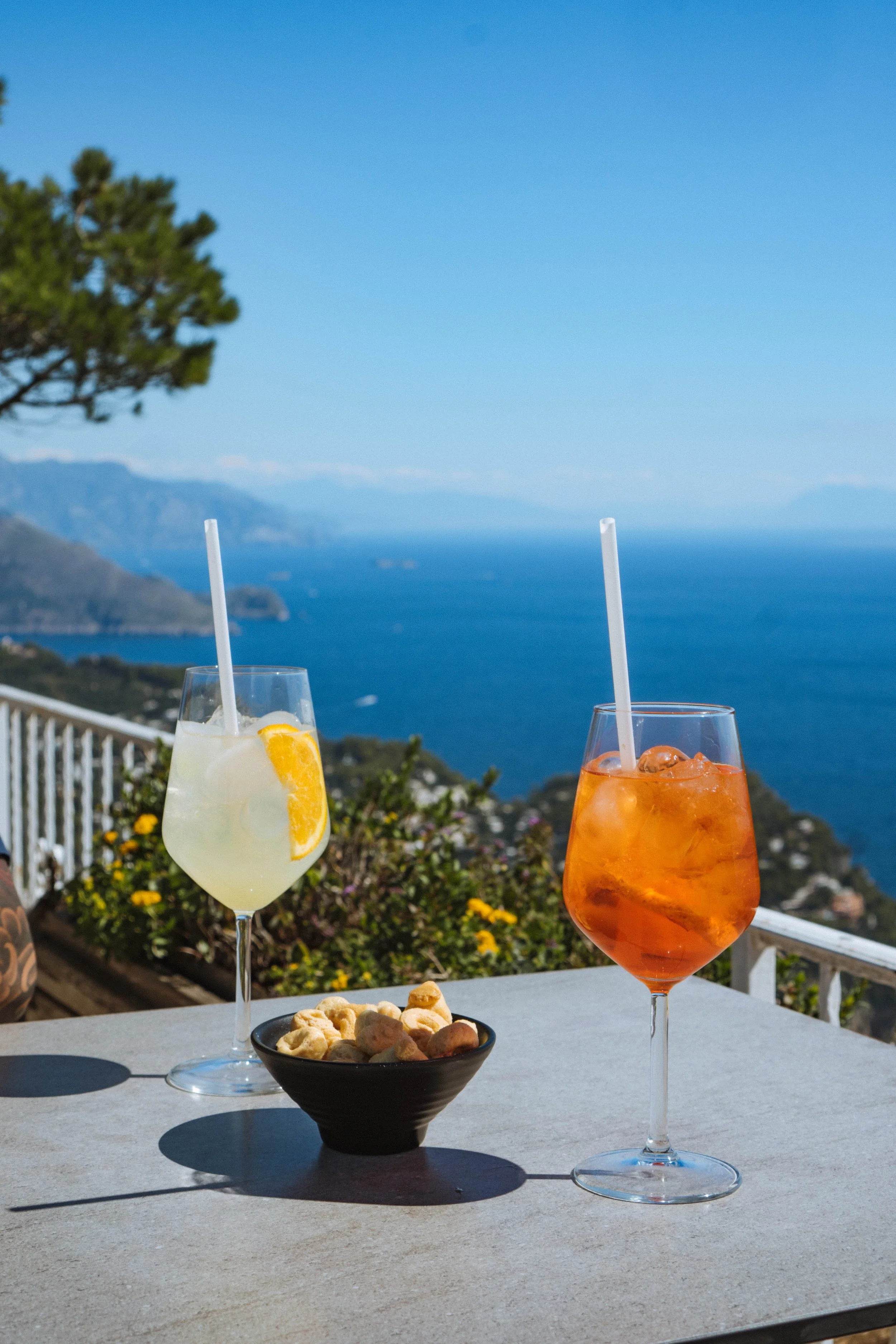 Two glasses of colorful cocktails with straws on a table, a bowl of snacks, and a scenic view of the ocean, mountains, and blue sky in the background.
