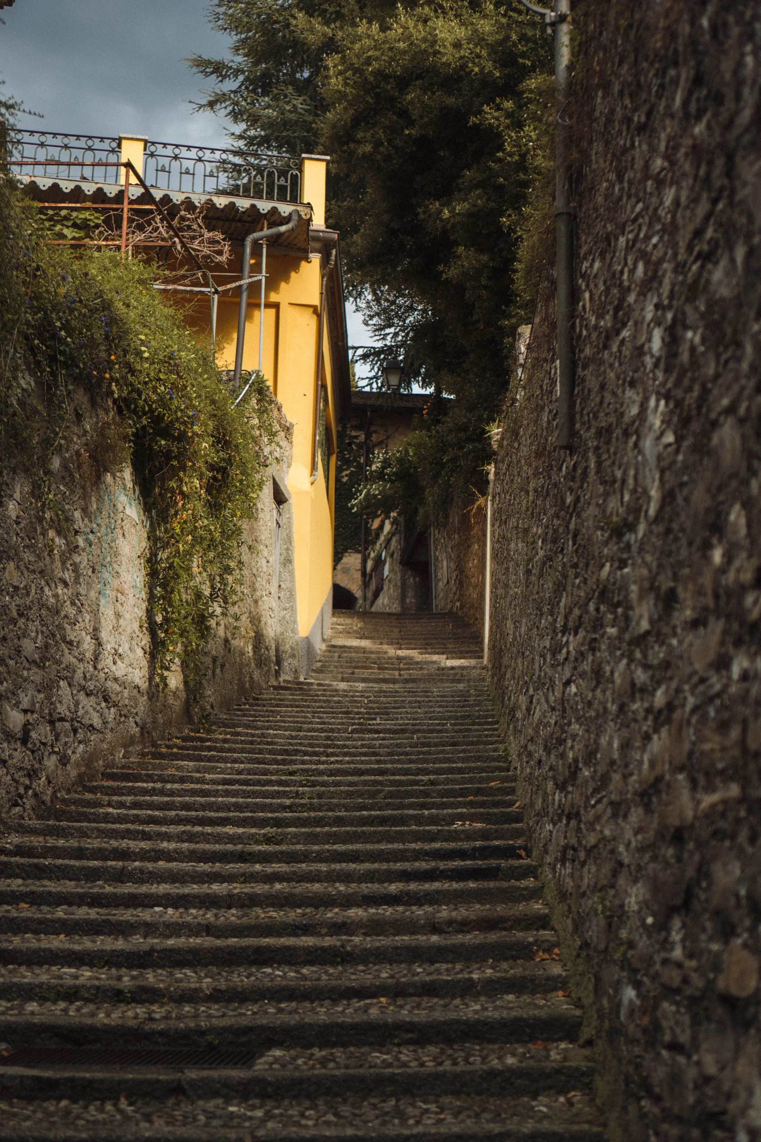 Stone stairs leading uphill between stone walls with greenery on the left and a yellow building with a balcony at the top.