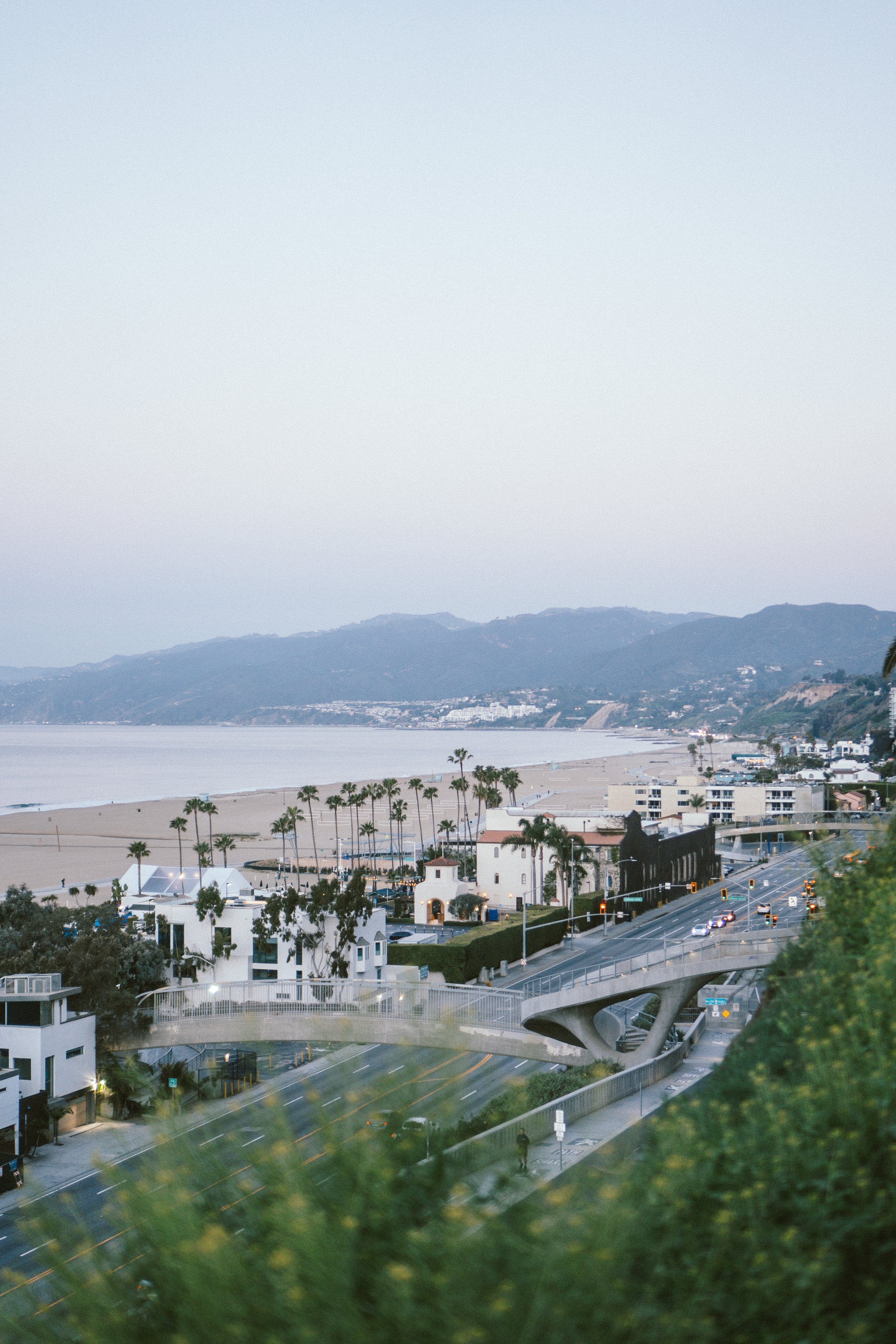 A coastal cityscape with high-rise buildings, palm trees, a beach, and mountains in the background, taken during the early evening.