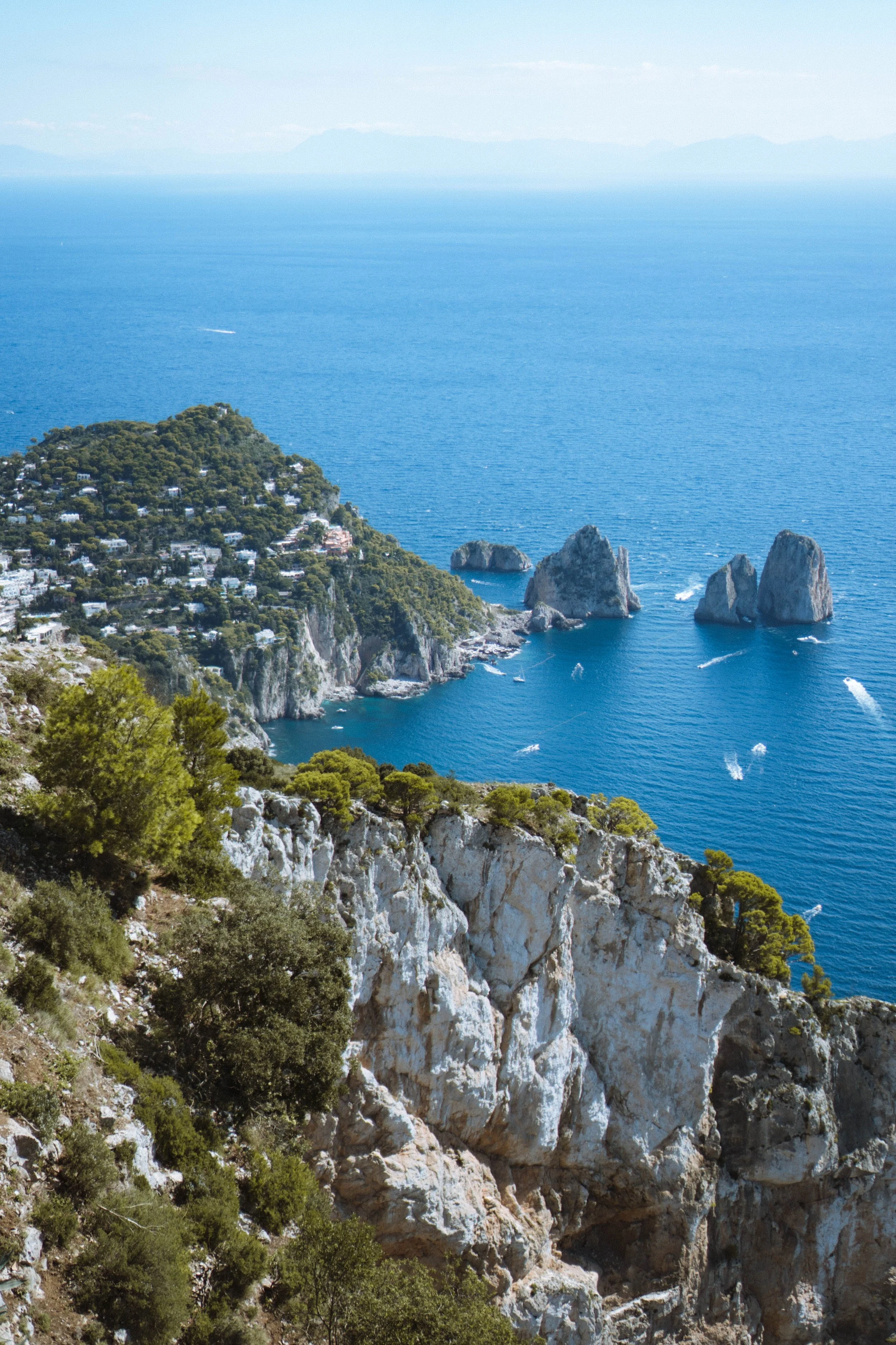 A coastal landscape featuring a green hill with houses, rocky cliffs, and the blue ocean with three large rocks near the shoreline. The water extends to the horizon where faint mountains can be seen in the distance.