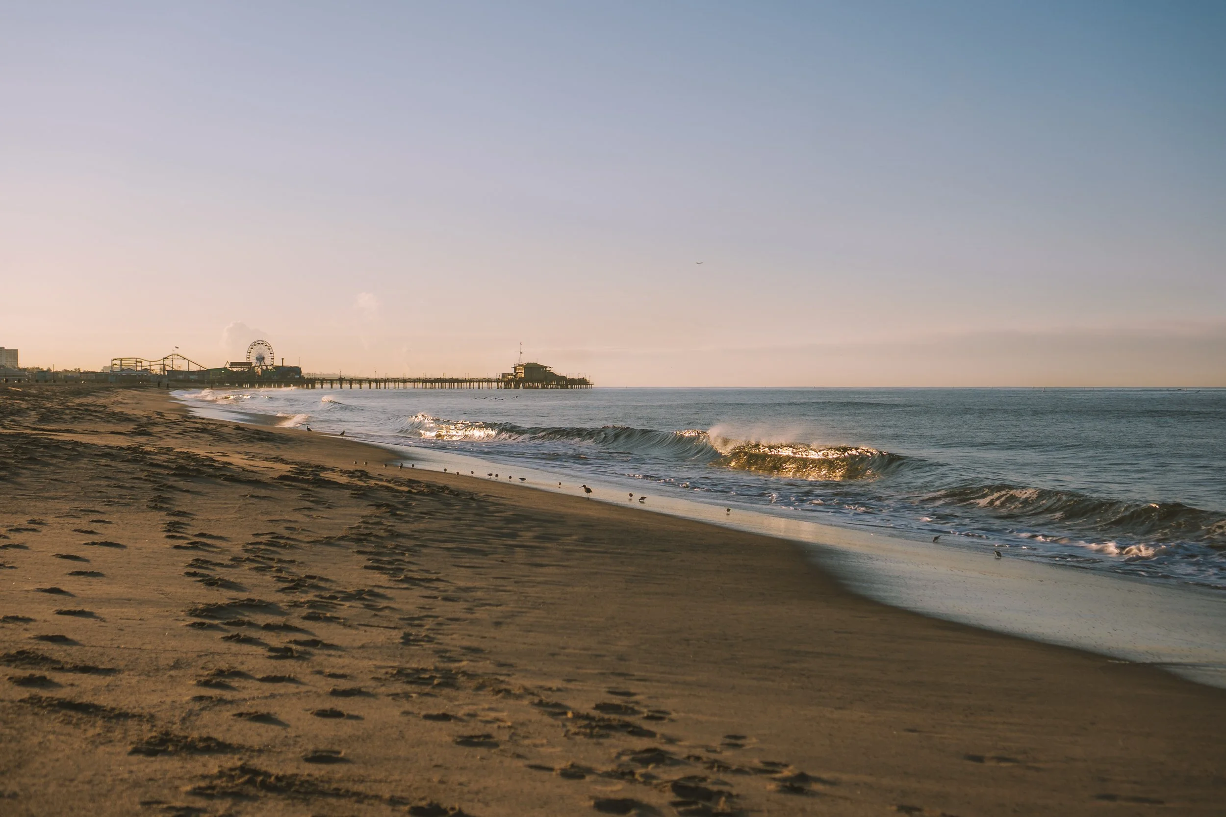 Sunset over a sandy beach with waves, a pier extending into the ocean, and a background with a Ferris wheel and amusement park rides.