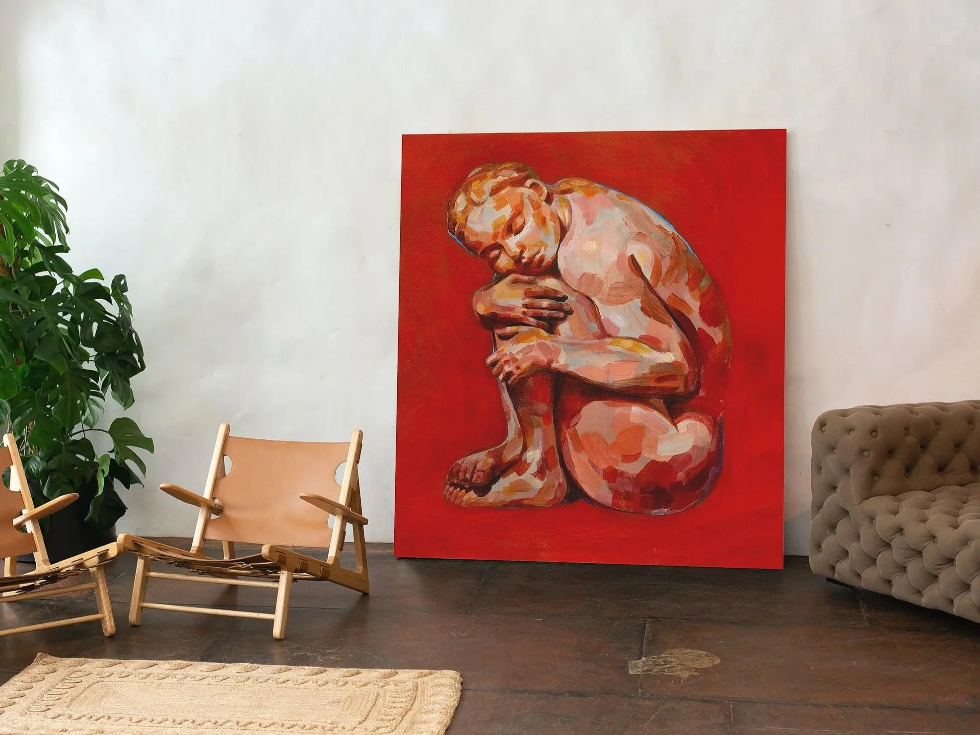 Interior of an art gallery with a large red canvas painting of a contemplative nude man sitting and hugging his knees on a wall, two wooden lounge chairs with fabric seats, a beige tufted sofa, a green potted plant, and a light-colored woven rug on a wooden floor.