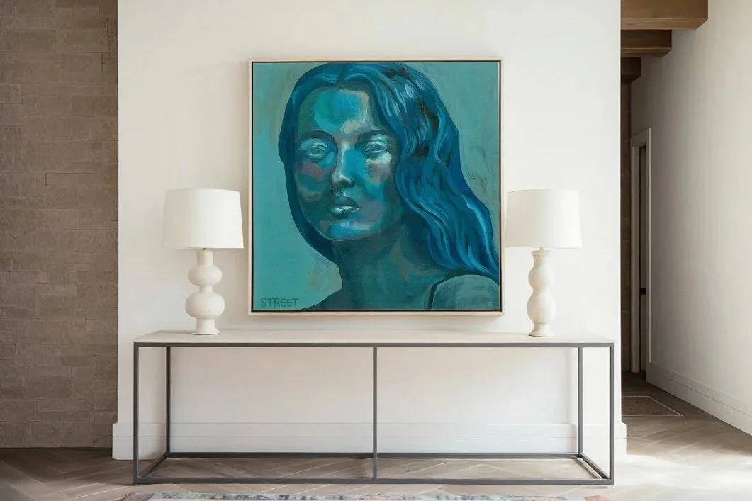 A painting of a woman's face in shades of blue hangs on a white wall. The painting is flanked by two white table lamps on a minimalist console table.