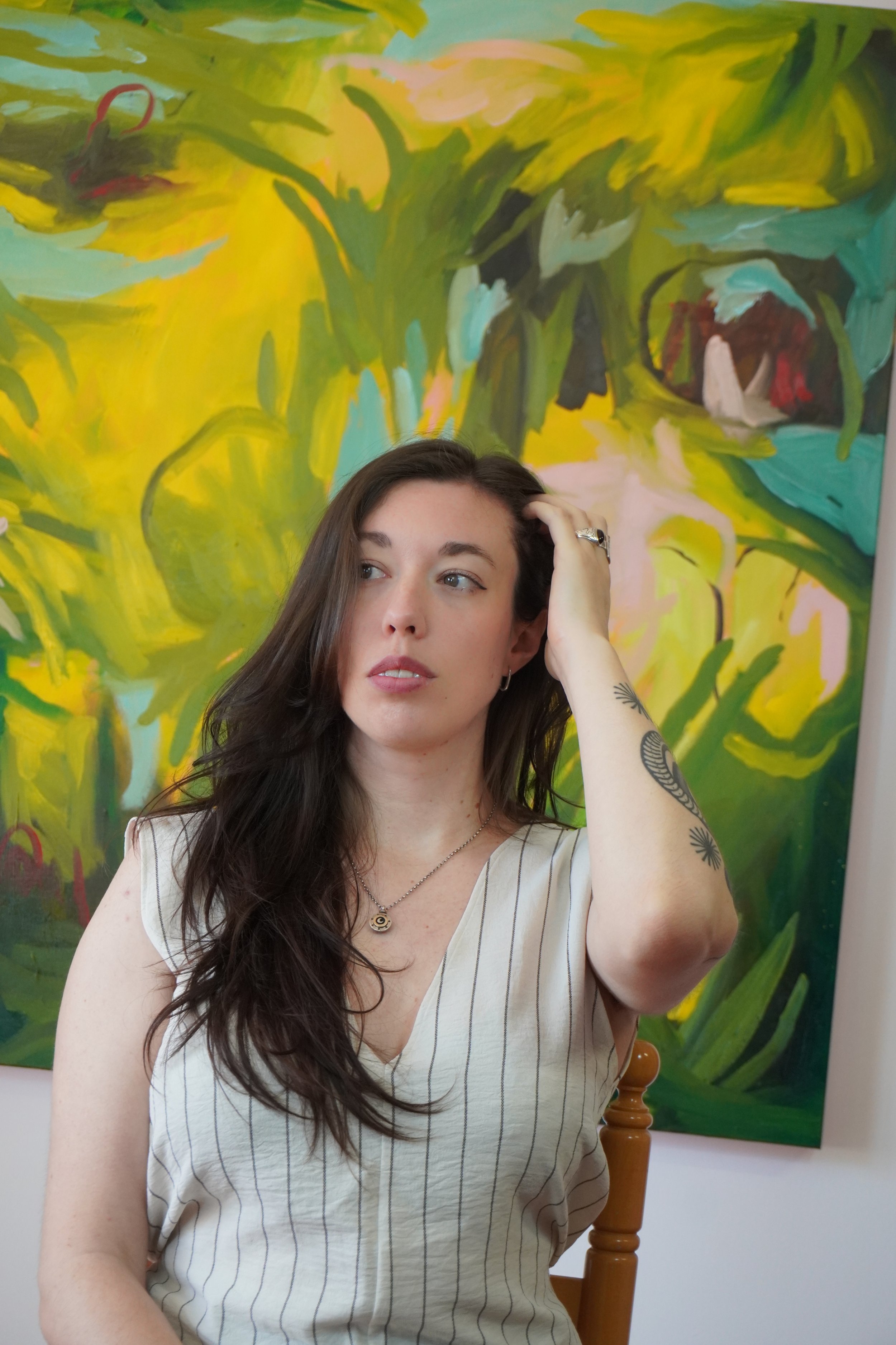 A young woman with long dark hair and tattoos on her arm, sitting in front of an abstract colorful painting, wearing a sleeveless striped top and a necklace, touching her hair while looking to the side.