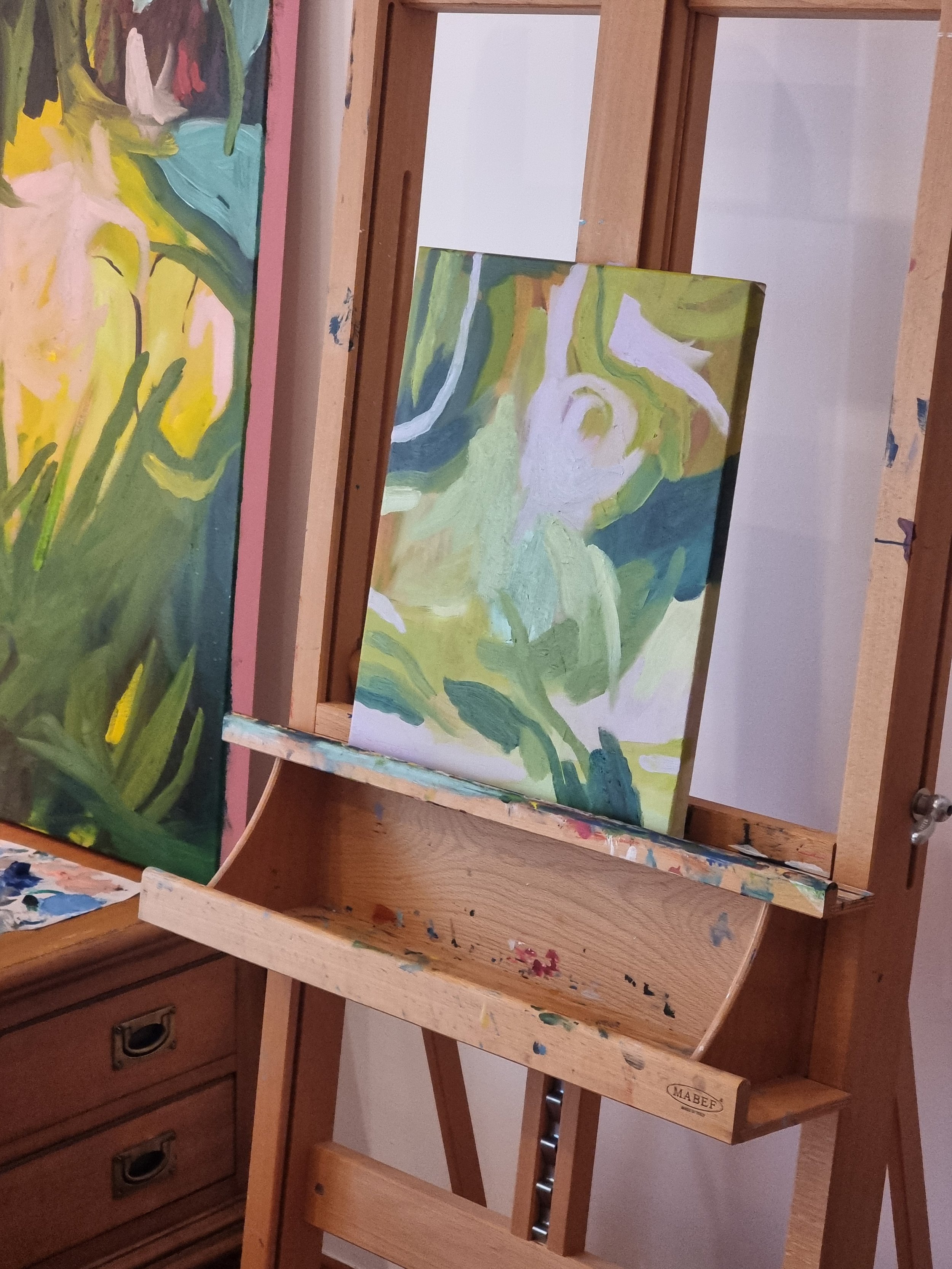 An abstract painting in progress on an easel, with green, white, and blue brushstrokes, featuring floral or leafy shapes.